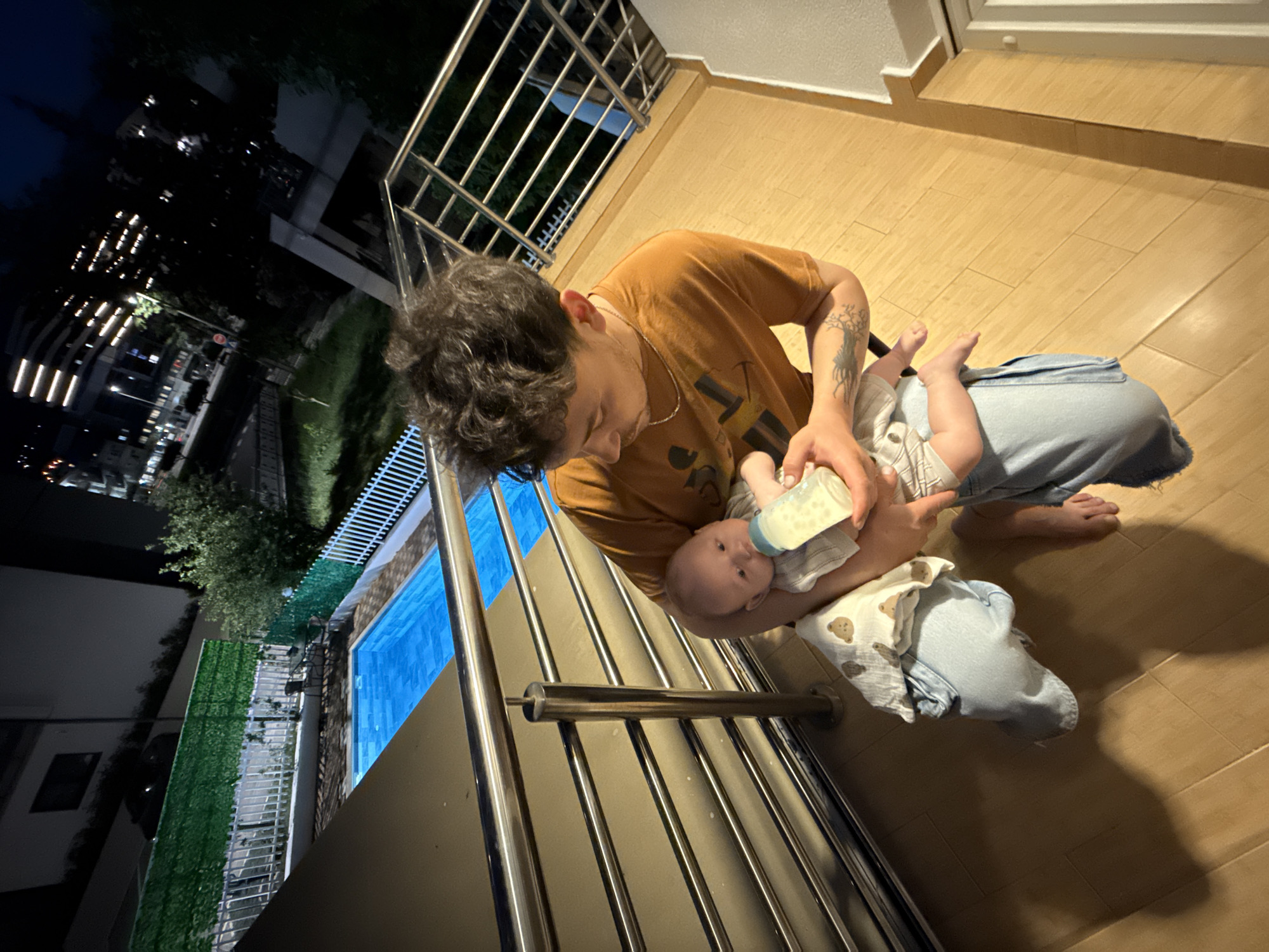 The image presents an overhead perspective of a man feeding a baby on a balcony at night. The man, with dark curly hair, is wearing an orange-brown t-shirt, light-colored pants, and is barefoot. He holds a baby bottle to the mouth of an infant cradled in his arm. The baby is dressed in a light-colored onesie, and a white blanket with a teddy bear pattern is draped over its legs. The man has tattoos on his left arm, one resembling a tree or plant.
The scene is set on a balcony with light-colored tiled flooring and prominent metal railings. To the man's left, a bright blue swimming pool is visible. Beyond the pool, there is a stretch of green landscaping, fences, and trees. The background reveals a dark nighttime city skyline, with numerous illuminated windows and architectural lights of buildings. A street with faint streetlights and a potential stop sign can also be seen. The dark sky confirms it is nighttime. The moment appears tender and calm, highlighting a caring interaction between the man and the baby in Bar, Montenegro. No clear brand text or prominent lettering is visible on any objects or clothing.