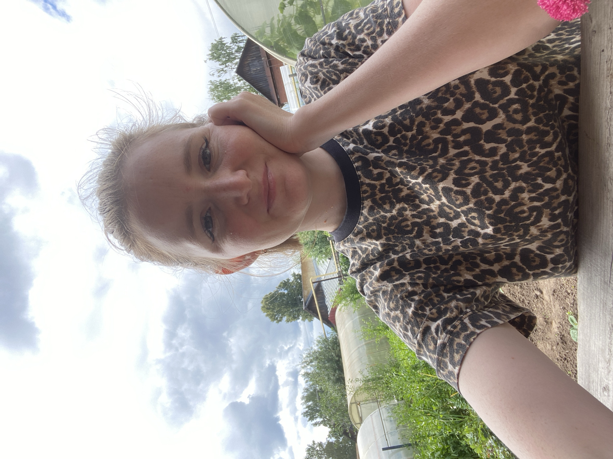 This is a selfie taken by a young woman outdoors. She is wearing a leopard-print t-shirt and has her hand resting on her cheek, with a slight smile. Her blonde hair is slightly windswept. The background shows a cloudy sky, green trees, and what appears to be a rural or garden setting with some sort of industrial or agricultural structures, possibly greenhouses or water tanks. The overall mood is casual and relaxed. It appears to be daytime, with overcast skies. There is no visible text in the image.