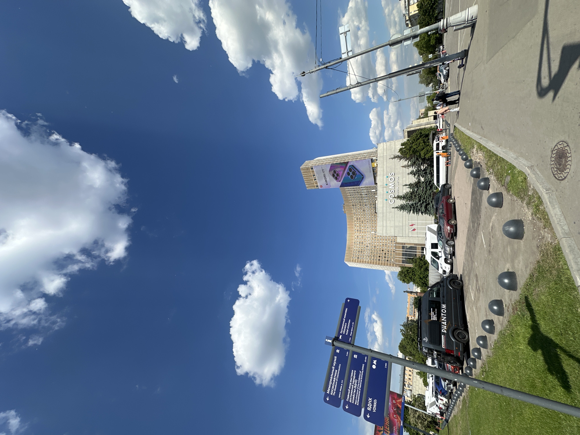 The image depicts a bright, sunny day in an urban setting, likely Moscow, Russia, given the visible text and provided context. The sky dominates the upper portion of the frame, a vibrant blue with scattered white cumulus clouds. Sunlight casts sharp shadows, indicating midday.
In the mid-ground, a large building with "COSMOS MOSCOW HOTEL" emblazoned on its facade stands prominently. Adjacent to it, another tall building features a large billboard advertising a "XIAOMI 11T" smartphone. Conifer trees are visible at the base of the Cosmos hotel.
The lower half of the image shows a sidewalk and a street running along the right side. A grassy verge separates the sidewalk from the road, lined with grey, dome-shaped bollards. Several vehicles are parked on the street, including a distinctive black van with "PHANTOM" written on its side, a white van/bus, and other cars. A few pedestrians are visible on the sidewalk in the distance.
On the left side, a multi-directional signpost with blue panels and white Cyrillic and English text provides directions to local landmarks. These signs point towards "The Museum of Cosmonautics," "Main Entrance of the Exhibition of Achievements of National Economy (VDNKh)", and the "Hotel «Cosmos»". Other visible text includes "ЦЕНА" (price) and various time indications like "10 min" and "2 min" associated with the directions. Infrastructure elements like streetlights, utility poles, and manhole covers are also present. The scene captures a typical, busy urban environment under clear skies.