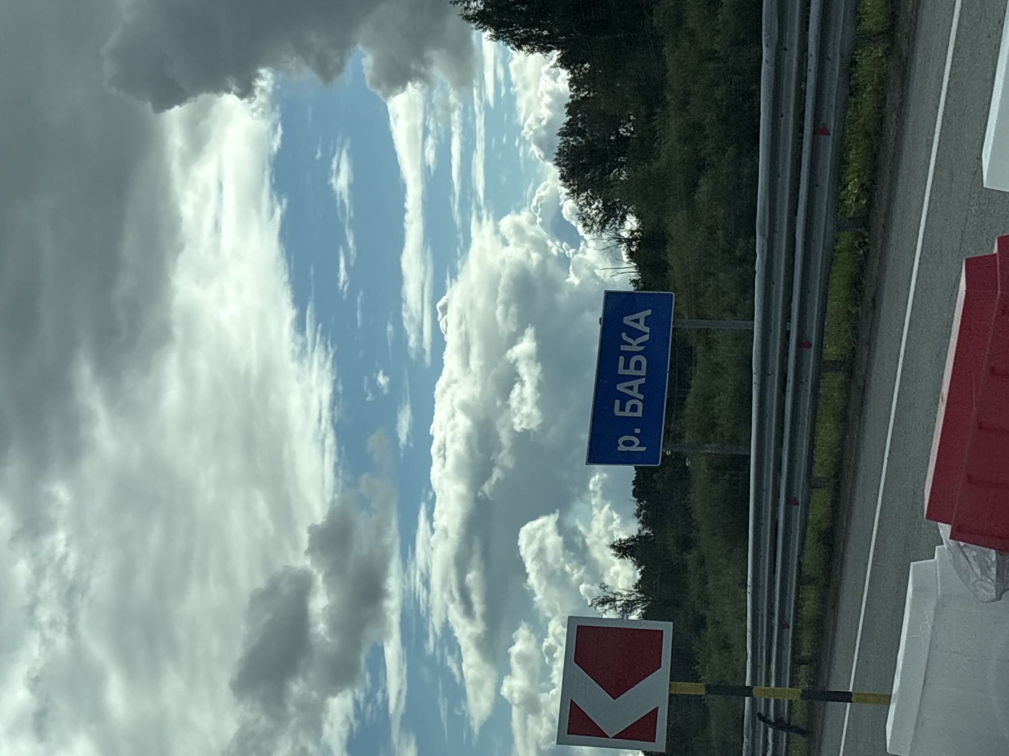 A photograph captures a roadside view under a partly cloudy sky. In the foreground, a highway barrier is visible, constructed of white and red plastic barriers. A metal guardrail runs along the edge of the road. To the left of the road, a sign with a red and white chevron indicates a road direction. Further to the right, a blue sign displays text in Cyrillic script, reading "р. БАБКА," which translates to "River Babka." The background consists of a dense line of green trees and bushes. The sky above is a mix of blue patches and large, dramatic white and gray clouds. The scene is set in the vicinity of Yanychi, Russia, specifically near the Babka River.