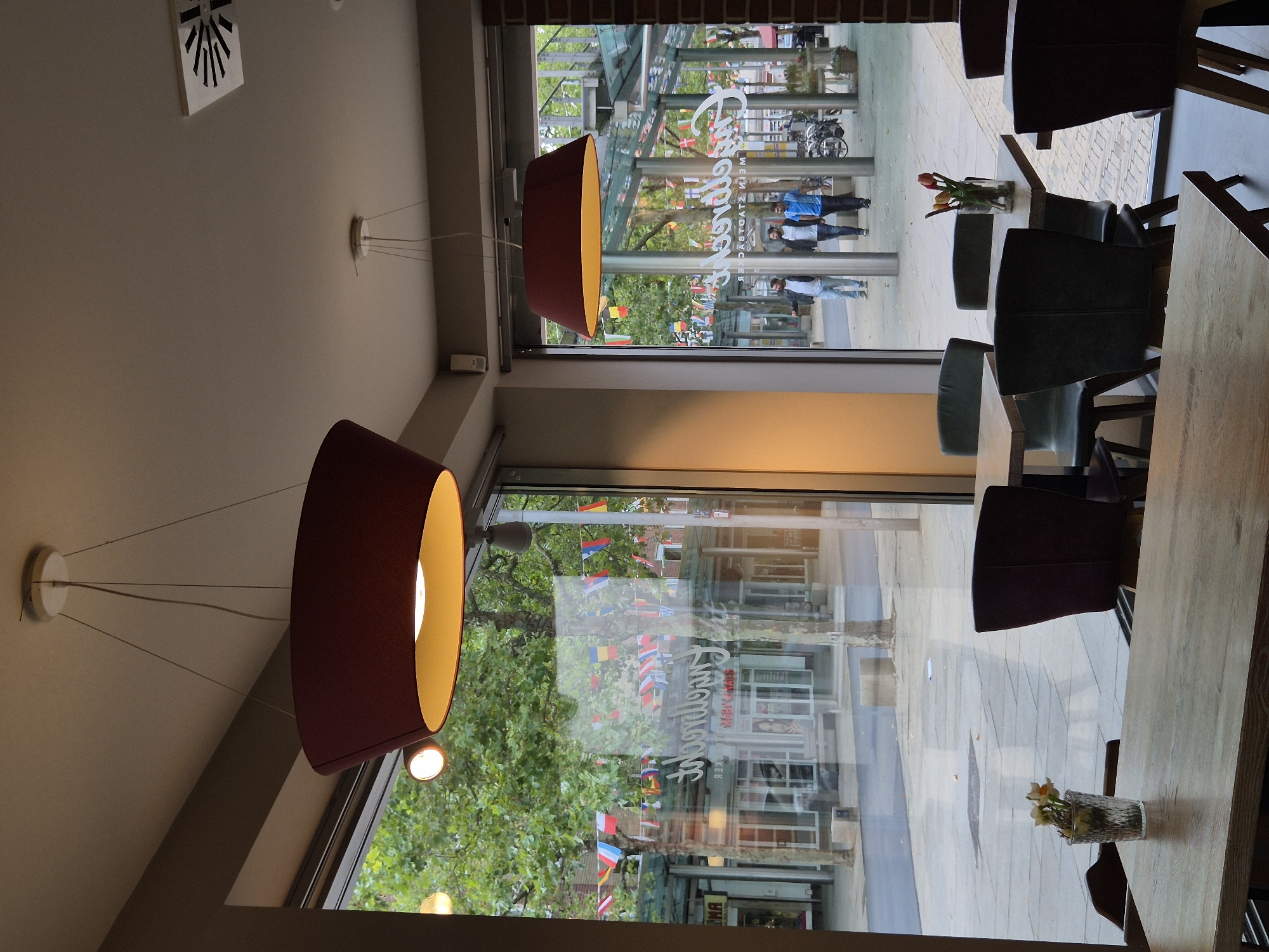 The image depicts the interior of a cafe or restaurant in Bremerhaven, Germany, with a large window offering a view of a street scene. Two large pendant lights hang from the ceiling, illuminating the dining area. Several tables with chairs are arranged inside, and small floral arrangements are visible on some of the tables. Through the window, a street with trees, shops, and flags is visible. Three men are walking on the street outside. A sign in the window, which is reversed due to reflection, reads "Freiraum Stadtbäckerei".