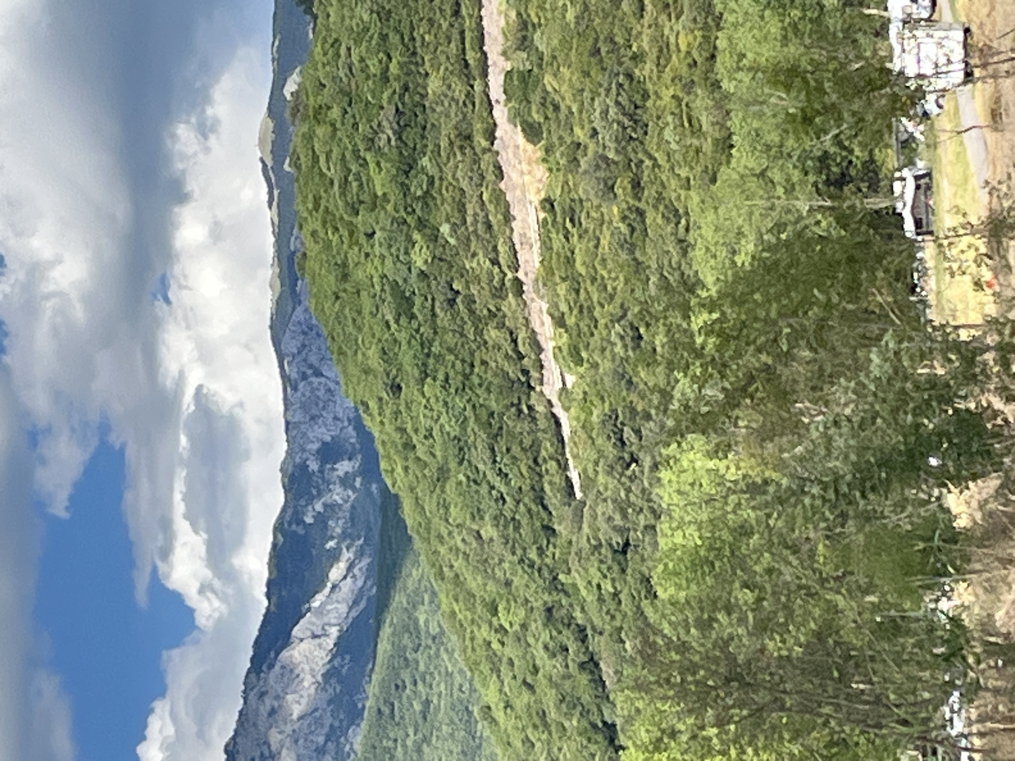 The image shows a mountainous landscape with lush green vegetation covering the slopes. In the foreground, there are trees and a campsite with several RVs and cars. The background features rugged, rocky mountains partially covered in snow, under a cloudy sky with patches of blue. The scene depicts a natural setting with recreational activity. The location is Prijevor, Montenegro.