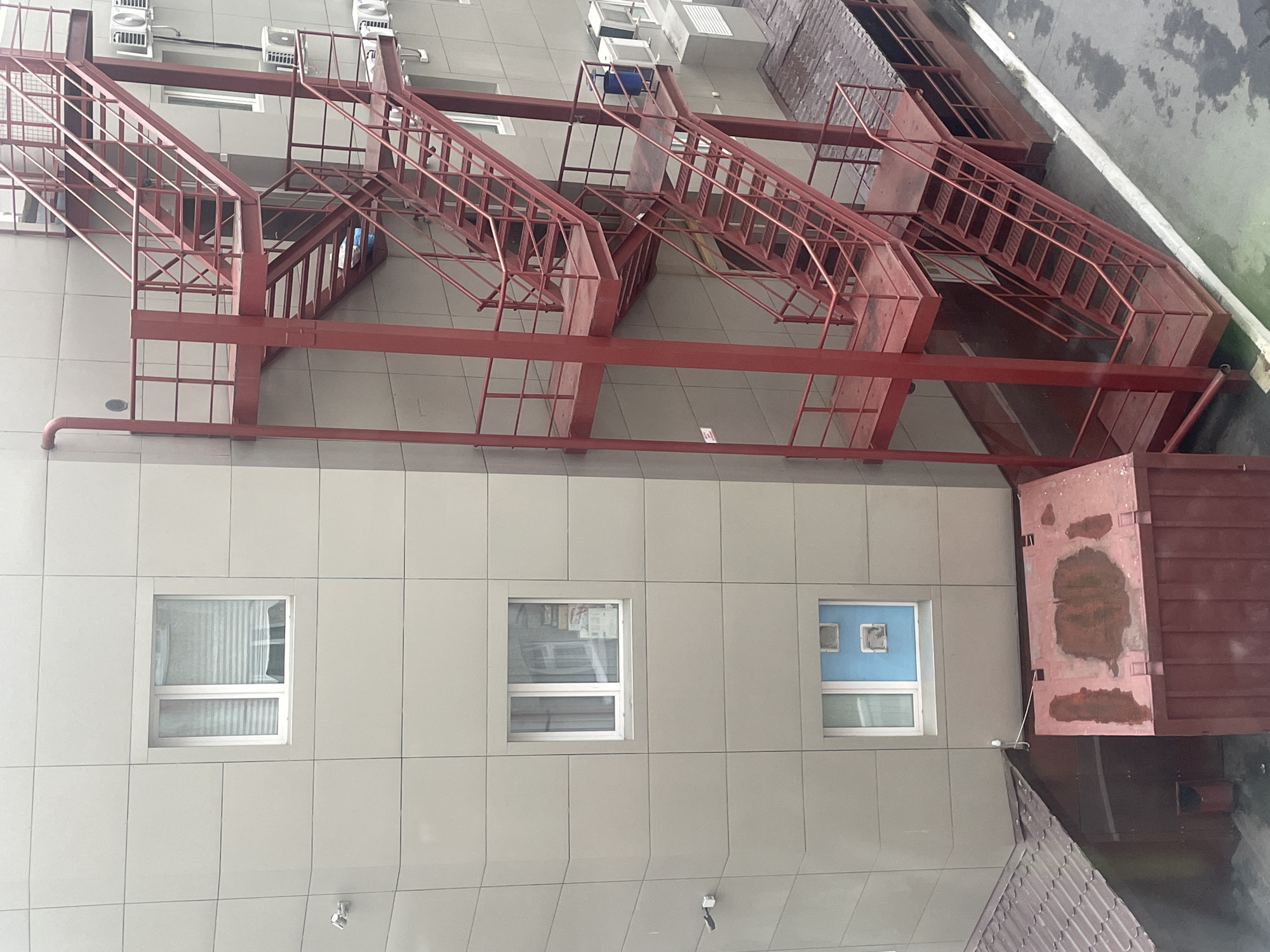 The image depicts the exterior of a building in Surgut, Russia, featuring a prominent red metal fire escape. The building has light-colored paneled walls with several windows. The fire escape consists of multiple flights of stairs and platforms, painted a vibrant red, attached to the side of the building. Below the fire escape, there is a red metal dumpster or storage container. To the left of the dumpster, a section of a brown tiled roof is visible, and next to it, a red trash receptacle. The ground appears to be asphalt or concrete. Air conditioning units are mounted on the building's exterior. There are no people visible in the image. The overall scene suggests an urban industrial or commercial setting.