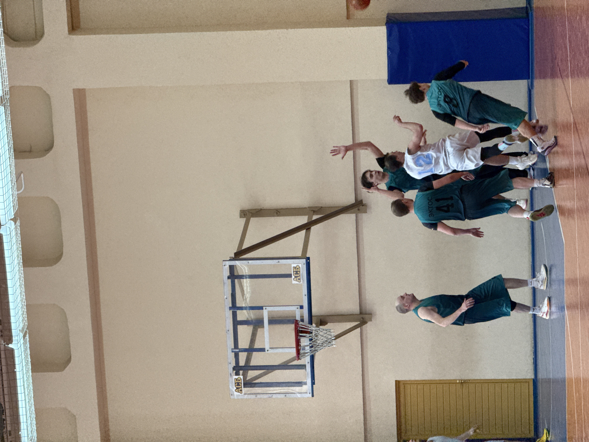 A basketball game is in progress. The players wear uniforms, and the basketball hoop is attached to the wall. The city is Chaikovsky, Russia.