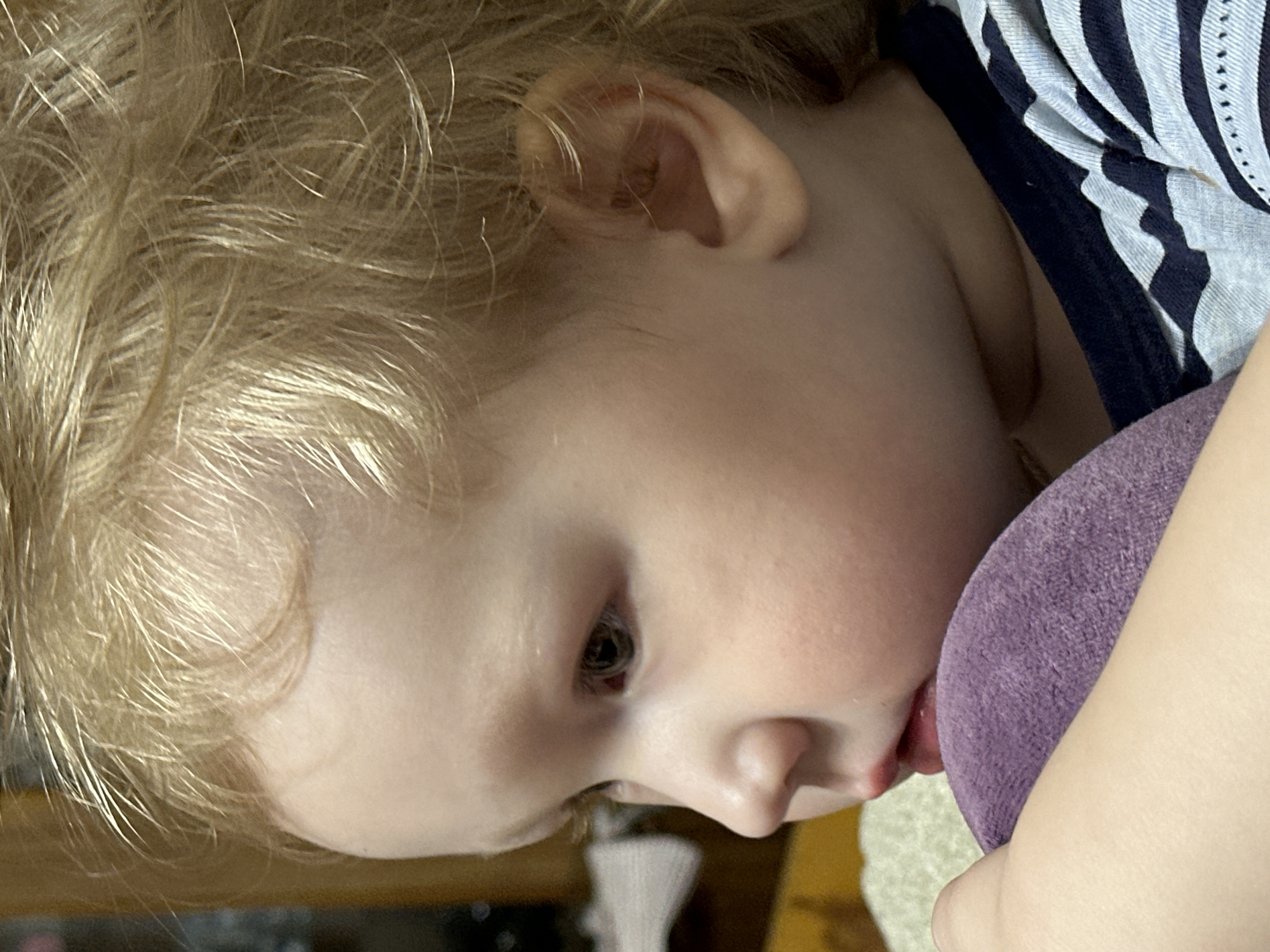 A close-up, rotated image features a young child with blond hair resting their face against a purple object. The child's face is partially visible, showing a closed eye, nose, and a portion of the mouth. The child is wearing a striped shirt. The location is not evident in the image.