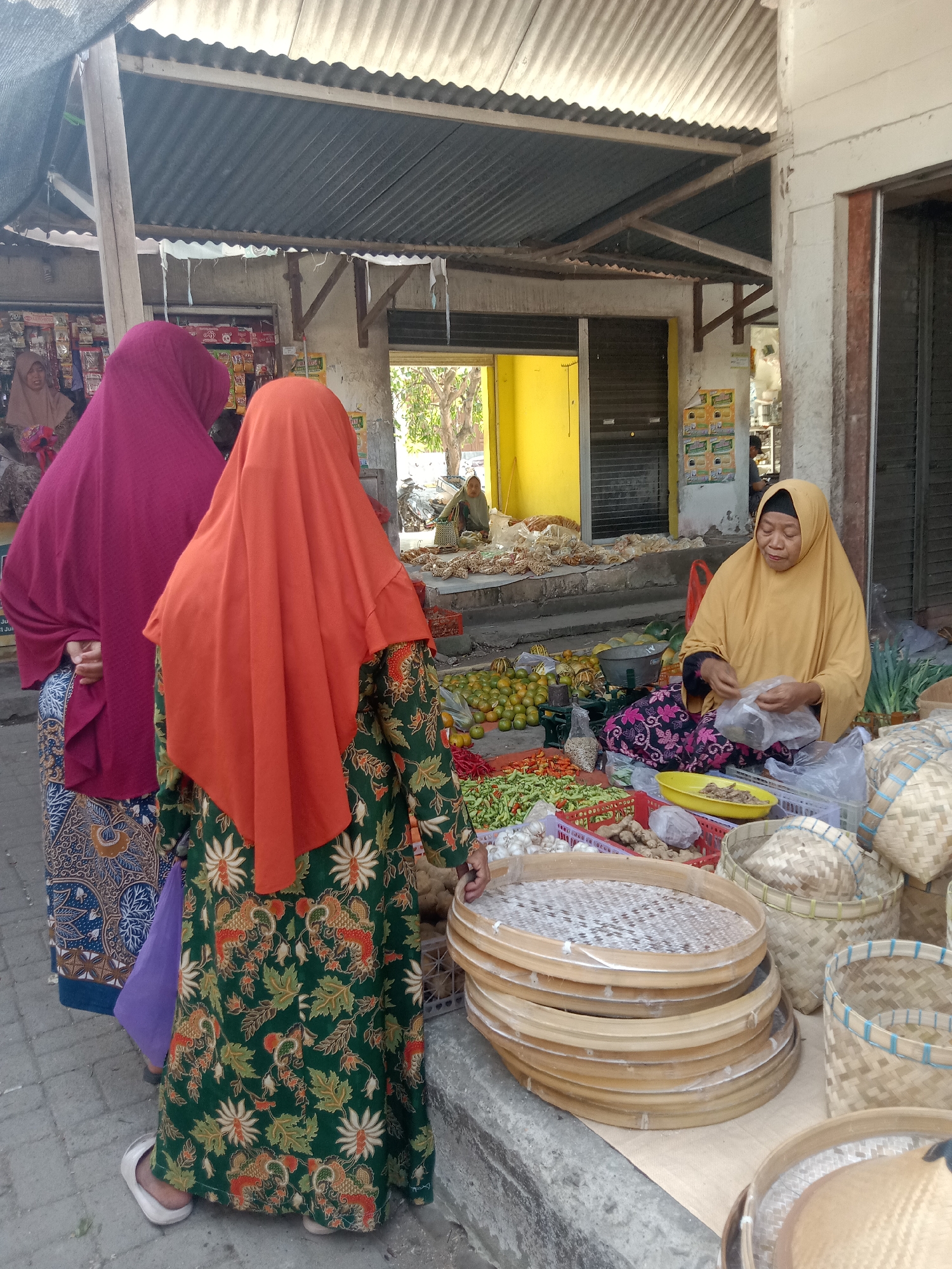 A bustling outdoor market scene unfolds in Klepek, Indonesia. The foreground is dominated by a stall laden with fresh produce and woven baskets. Several people are present, including a vendor in a yellow hijab, seated and busily bagging goods. Three women, two of whom are wearing vibrant hijabs and patterned clothing, are interacting with the vendor and examining the merchandise. In the background, a building with a corrugated metal roof and yellow walls suggests the market's permanent structures. The overall atmosphere is one of lively commerce and everyday life in a Indonesian marketplace.