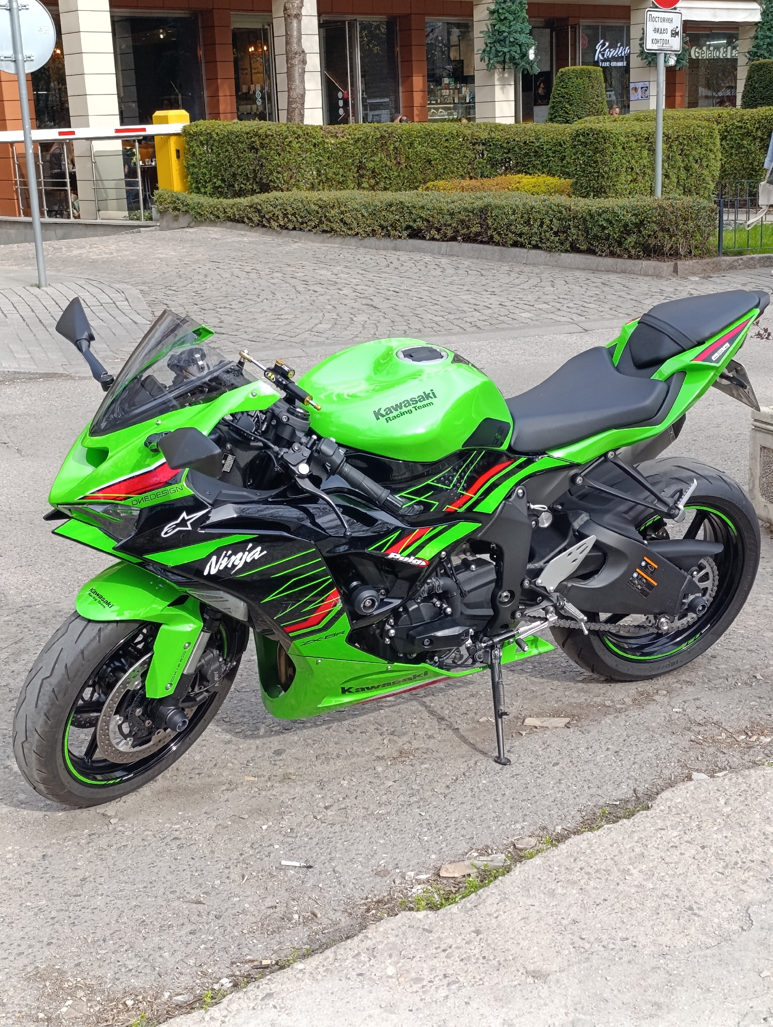 The image shows a Kawasaki Ninja ZX-6R motorcycle parked on a paved surface. The motorcycle is predominantly green and black. It features "Ninja" graphics on the side and branding for "Kawasaki Racing Team."
The motorcycle is positioned on a city street in Sofia, Bulgaria. In the background, there is a hedge and a building with storefronts. There is signage, including one that reads "Постоянен Видео контрол" (Permanent Video Control) and another that mentions "Gelato & La". The environment suggests a commercial or mixed-use area in Sofia.