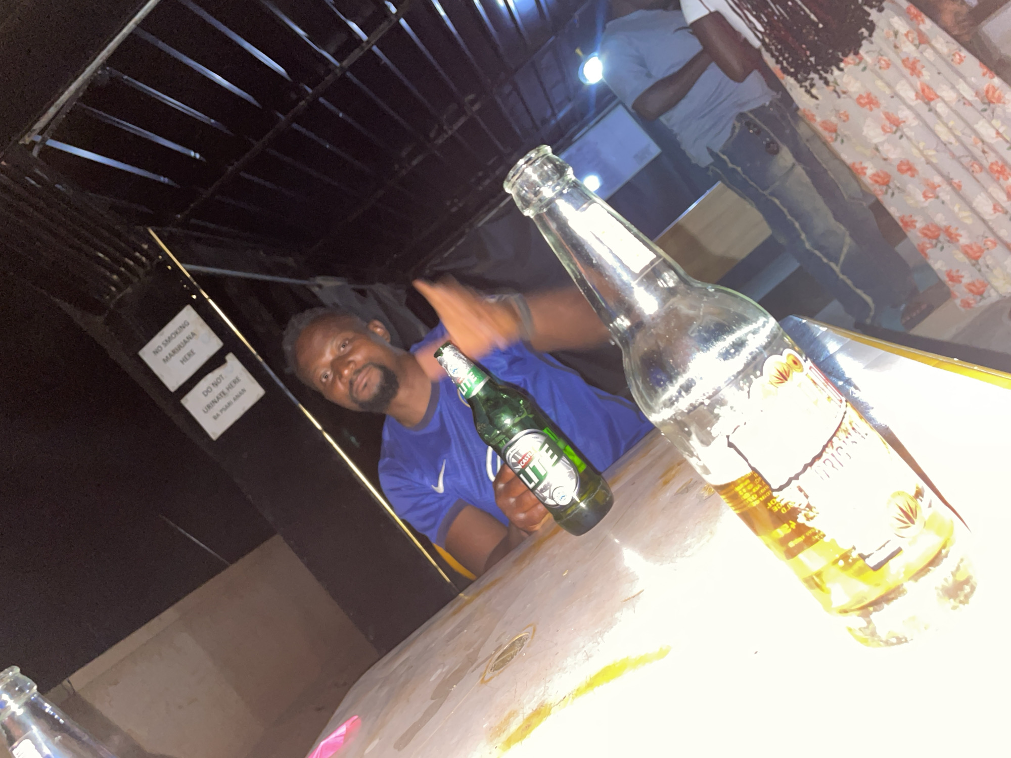 The image depicts a man seated at a table with two beer bottles, one Castle Lite and the other Star. The table has a faded or worn appearance. A blurred figure stands in the background, wearing jeans and a light-colored shirt. Another individual, partially visible, is wearing a floral dress. Above the man, signs read "NO SMOKING MARIRIANA HERE" and "DO NOT URINATE HERE BA PSARI ANAN". The setting appears to be a bar or outdoor seating area with a dark, overhead structure of metal bars. Based on the signage with "Ba Psari Anan", the location is likely in or near Jos, Nigeria.