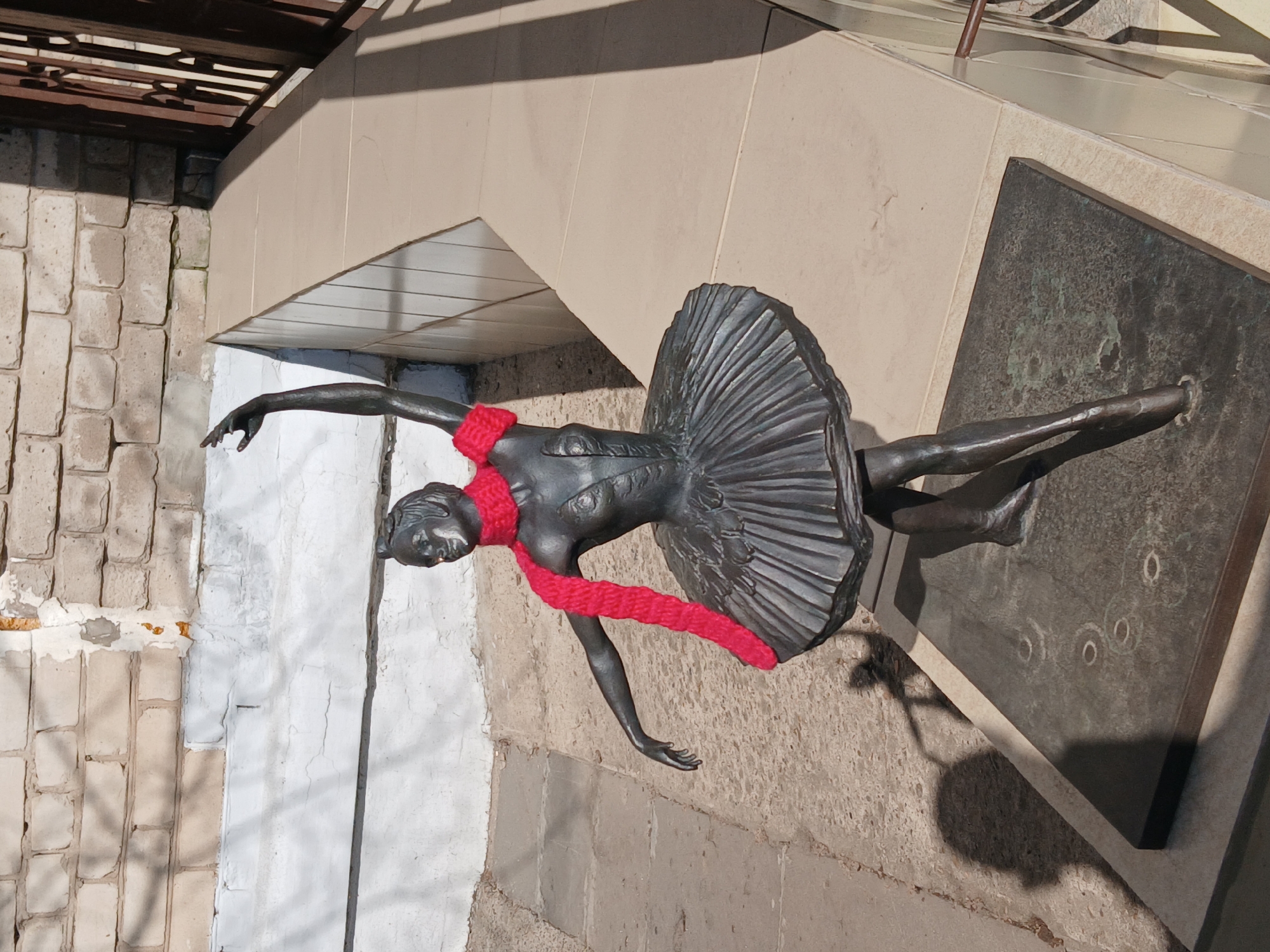 A bronze statue of a ballerina wearing a red scarf stands in the city of Chaikovsky, Russia. The statue is mounted on a concrete and stone base with a bronze plaque. The statue is in a ballet pose, with one leg extended and arms outstretched. The background consists of an arched beige and grey concrete wall with white brick to the left and shadows in the upper left corner.