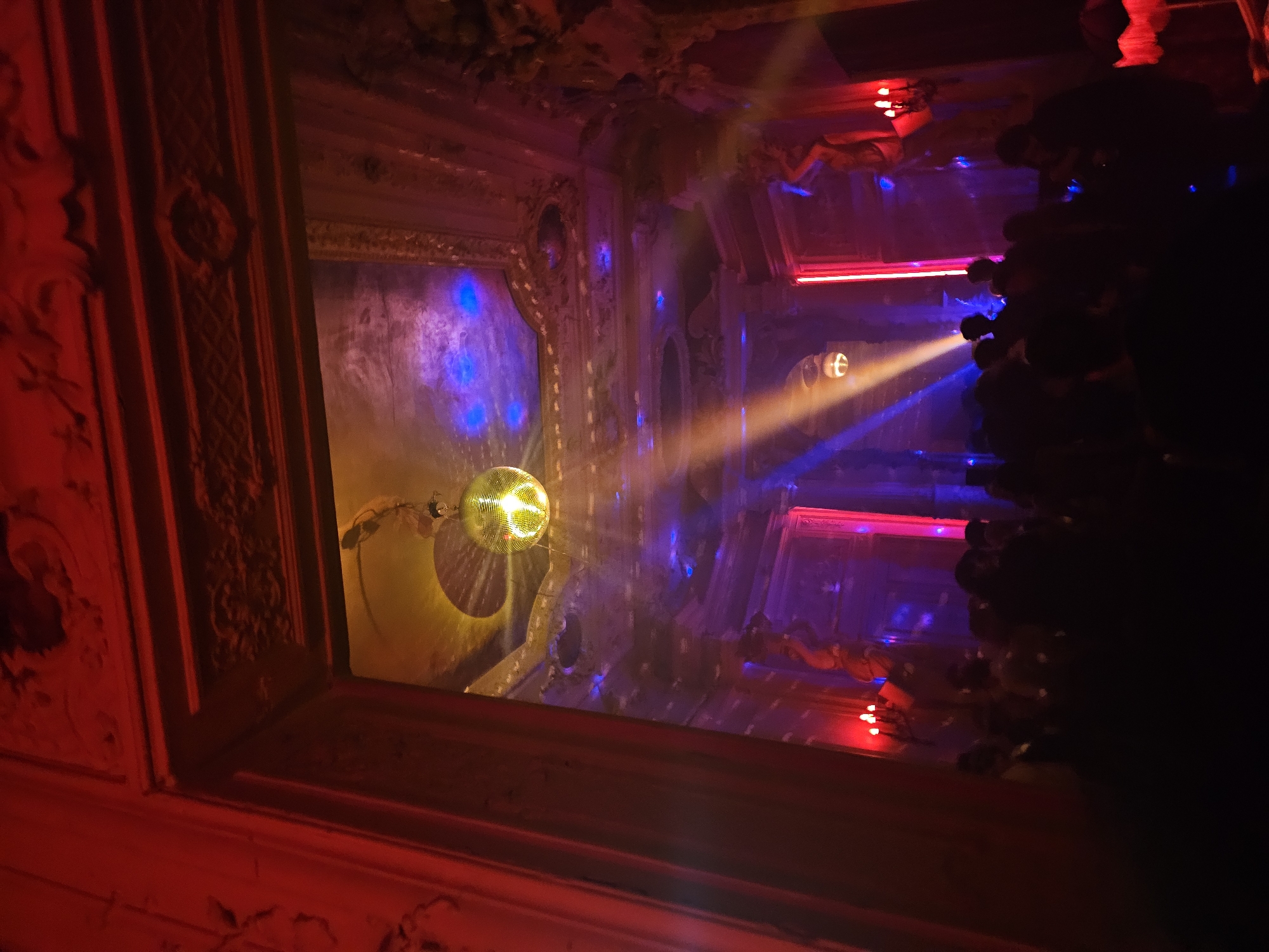 The image shows a reflection of a disco ball and stage lighting in a mirror, framed by an ornate red and gold-colored trim. The lighting includes blue, red, and yellow hues, creating a colorful and energetic atmosphere. The mirror reflects a space with additional decorative elements and lighting fixtures. The location is in Paris, France.