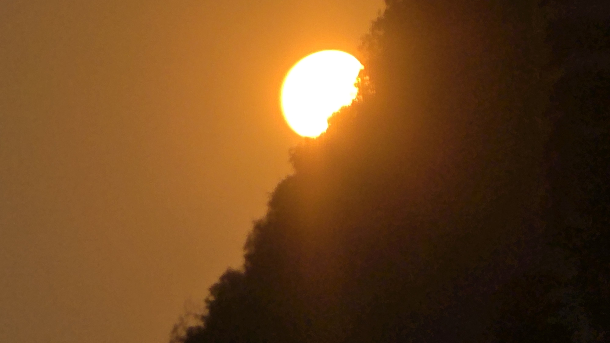 The image depicts a bright, yellowish-orange sun partially obscured by a dark, irregular mass, likely a mountain or large hill. The sky has a hazy, orange hue. The location is in the city of Bac Natural Heritage Area Trang, Vietnam.