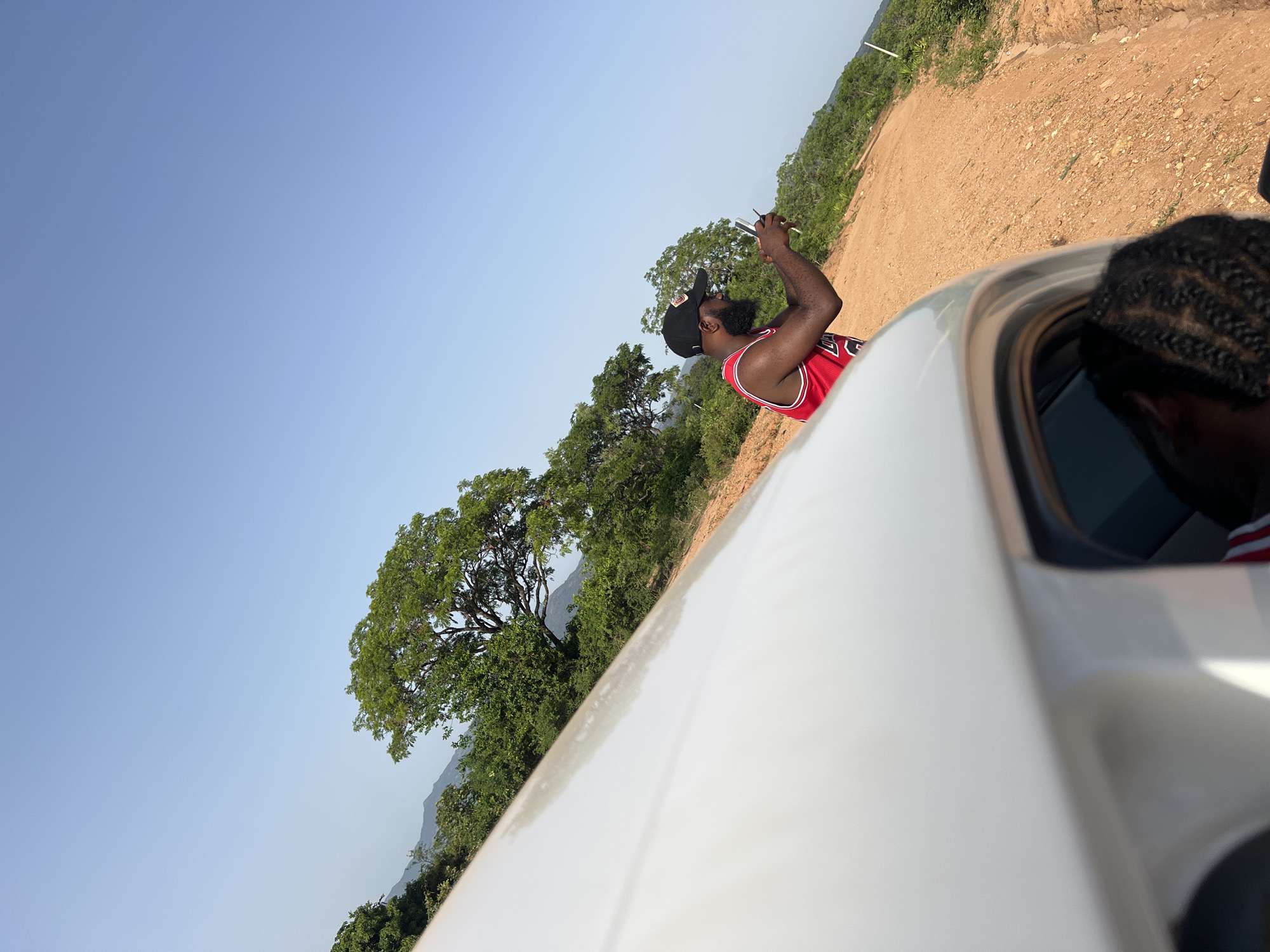 A man with a beard and wearing a red and white basketball jersey and a black baseball cap is seen outside of a car, holding a phone up to take a picture. Part of another person with braided hair is visible in the car window. The environment includes trees, a dirt road, and a clear blue sky. This appears to be in a rural area of Fobur, Nigeria.