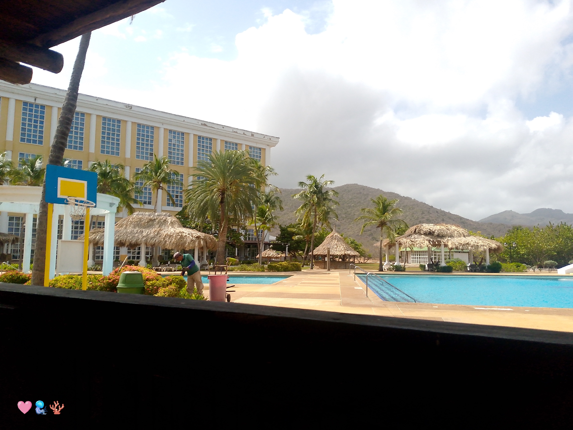 The image shows a view of a resort-style location in Pedro González, Venezuela. A large swimming pool with turquoise water is visible, along with thatched-roof cabanas. A multi-story yellow building with numerous windows is on the left. Palm trees and other tropical vegetation dot the landscape. Mountains are in the background. A man in a green shirt is working in the landscaping near a green trash can and a pink bin. The shot appears to be taken from an elevated, partially obstructed view, as indicated by the dark foreground. The sky is partly cloudy.