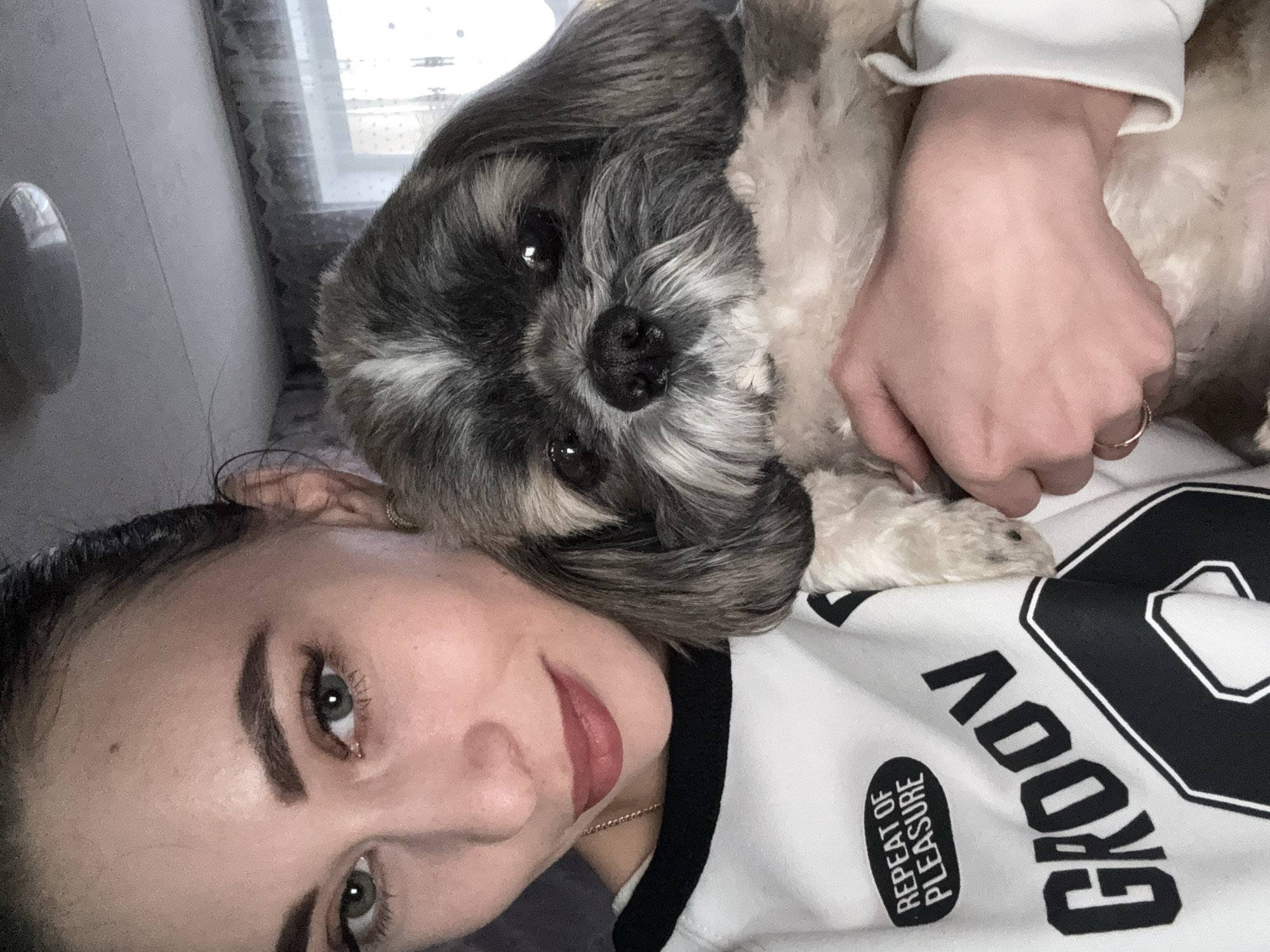 This close-up, rotated portrait captures a young woman and her dog in an indoor setting, likely in Chaikovsky, Russia. The woman is positioned on the left side of the frame, looking towards the camera with gentle, expressive green eyes and a subtle, warm smile. She has dark eyebrows and soft, naturally colored lips. Her dark hair is partially visible around her ear. On her right ring finger, she wears a simple gold ring. She is dressed in a white sweatshirt adorned with large black lettering, part of which reads "GROOV" (suggesting "GROOVY"), and a smaller black oval patch inscribed with the white text "REPEAT OF PLEASURE."
Cuddled close to the woman, resting its head near her neck and shoulder, is a small, fluffy dog, possibly a Shih Tzu. The dog features a charming mix of dark grey/black and white fur on its head and body, and looks directly at the camera with dark, engaging eyes and a prominent black nose.
The scene conveys a moment of affectionate companionship and peaceful interaction between the woman and her pet. The lighting in the image appears to be natural daylight, softly illuminating the subjects, with a window and curtains visible in the blurred background, suggesting a cozy domestic environment. The overall mood is one of warmth, comfort, and tenderness. The image is taken from a slightly high and tilted angle, indicative of a casual, perhaps self-portrait, style.
