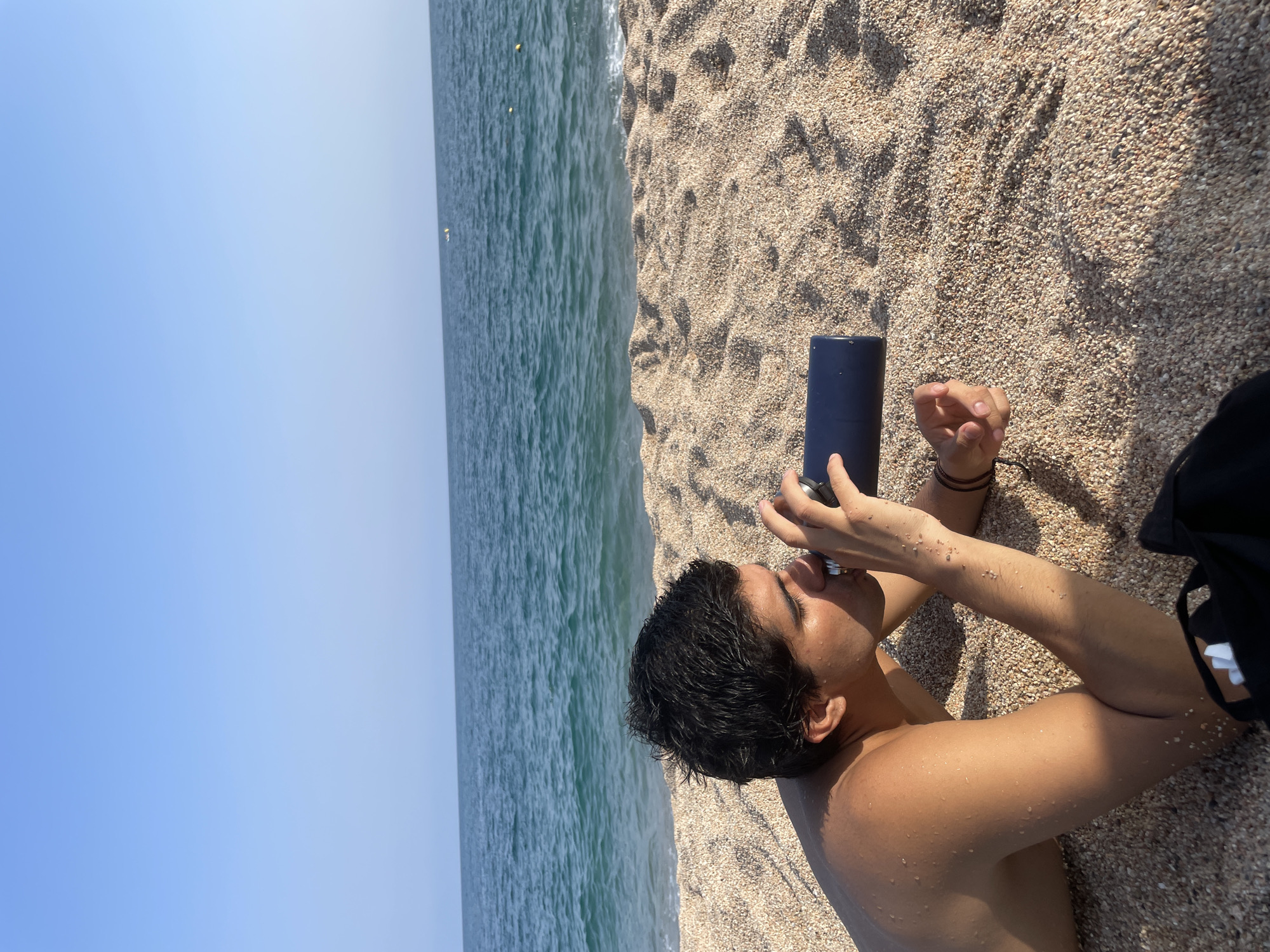 A person is lying on a sandy beach in Canet de Mar, Spain, drinking from a dark blue water bottle. The person is facing left, with their body on the sand and their head resting on their arms. The ocean is visible in the background, with waves gently lapping at the shore. The sky is clear and blue. The sand is light brown and speckled with small pebbles. A black bag is partially visible in the lower right corner of the image.