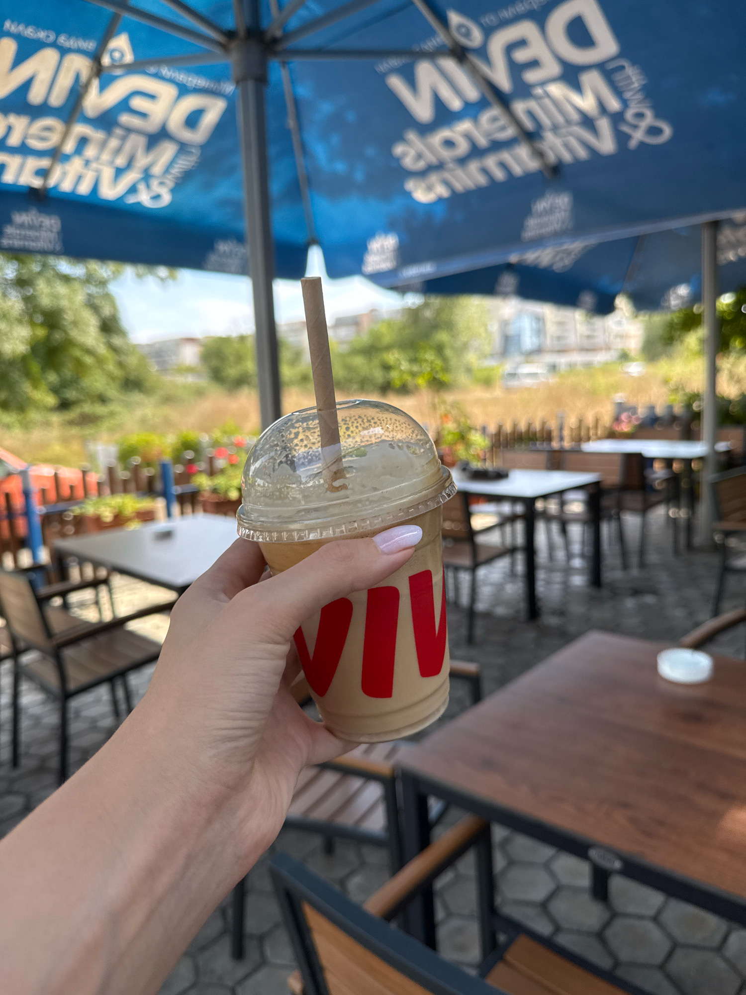 In an outdoor seating area, a hand holds a plastic cup of iced coffee topped with a dome lid and a paper straw. The cup has a red logo on it. The background shows tables and chairs, and a large blue umbrella with white lettering. Trees and buildings are visible in the distance.