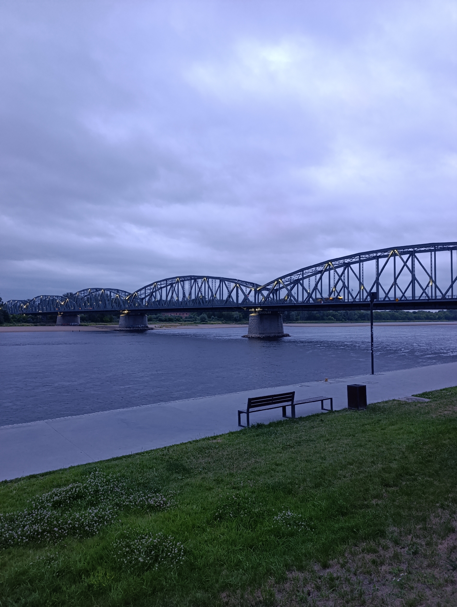 The image depicts the Toruń railway bridge, a prominent landmark in Toruń, Poland, spanning the Vistula River. The scene is captured during twilight, under a cloudy sky, casting a muted, blueish-grey light over the environment.
The bridge, a multi-span steel truss structure, is illuminated by numerous small lights along its sides, creating a warm contrast with the cool-toned sky and water. The river itself appears dark and slightly rippled, reflecting the subdued lighting.
In the foreground, a paved walkway runs parallel to the riverbank, which is covered in lush green grass. A simple wooden bench and a dark-colored trash receptacle are situated on the walkway, suggesting a public recreational area. Small white wildflowers are scattered amongst the grass. The overall atmosphere is serene and somewhat melancholic, characteristic of dusk in an urban setting. There are no people visible in the frame.