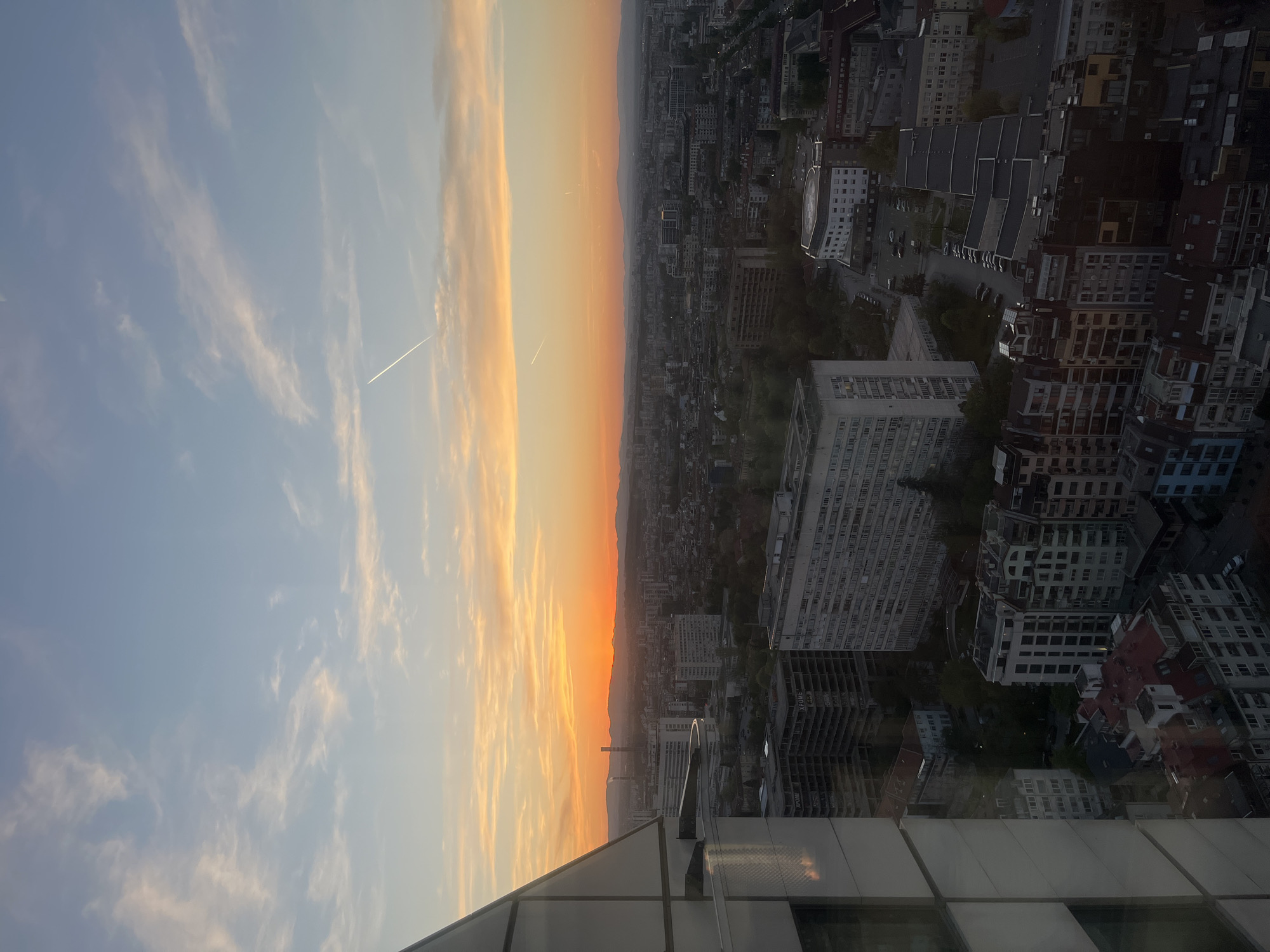 The image depicts a sunset over Sofia, Bulgaria, as viewed from a high vantage point within a modern building. The sky transitions from a light blue with wispy clouds to a vibrant orange and yellow gradient near the horizon. A contrail from an airplane is visible in the blue sky. Below the horizon, the urban landscape of Sofia extends into the distance, with buildings of varying sizes and architectural styles. There is no visible activity on the streets below. A foreground element shows the top and facade of the building from which the photo was taken, featuring reflective glass panels.
