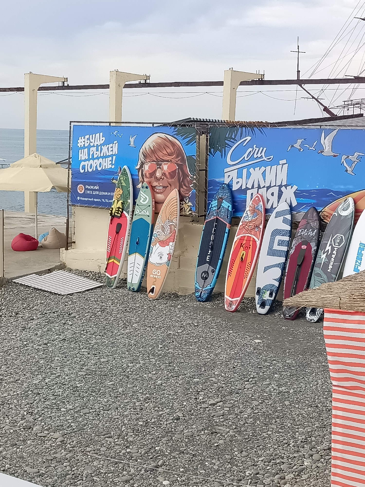 The image depicts a beachside scene in Sochi, Russia, with a row of stand-up paddleboards (SUPs) leaning against a wall. The SUPs feature various colorful designs and logos, including "GO BOARD," "DASHA," and "MAGNUM PRO." In the background, a large blue banner displays the Russian text "#будь нарыжей стороне!" (Be on the side of fun!) and an illustration of a person wearing sunglasses and a lei. The environment is characterized by a pebble beach, a partially visible tent or awning, and the sea in the distance. The sky is overcast, suggesting cloudy weather. There are no visible people actively engaged in activities within the frame, but the presence of beach equipment and the advertising banner implies a recreational or rental area. The overall impression is one of a watersports venue or a promotional display for such activities in Sochi.