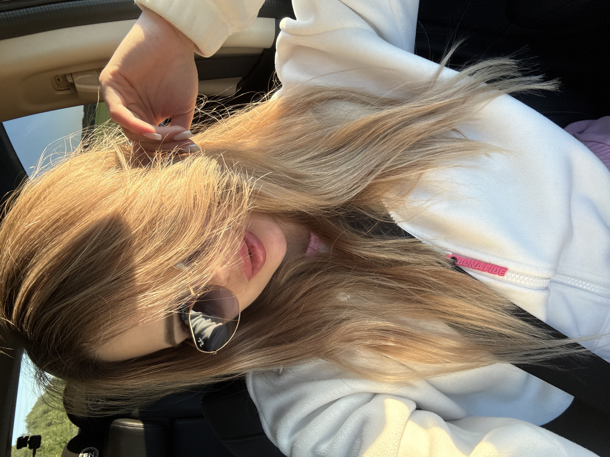 A woman with long blonde hair is sitting in a car, wearing sunglasses and a white jacket. Her hair is partially obscuring her face, and she is touching her hair with her right hand. The sun is shining, casting a warm glow on her hair and face. The background shows a glimpse of trees and a blue sky through the car window, suggesting an outdoor setting. The interior of the car is visible, with black seats and a partial view of the dashboard. A pink zipper pull with the word "BONAFIDE" is visible on her jacket.
