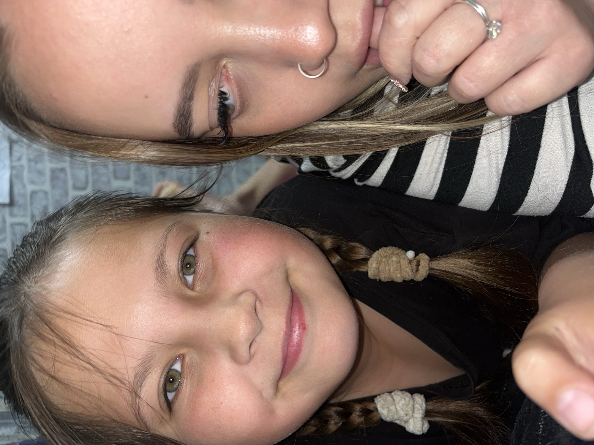 A close-up, tilted shot shows two females posing for the camera indoors. The upper figure is an adult with light skin, a nose ring, long eyelashes, and blonde hair with streaks. She is wearing a black and white striped top. Her hand is raised to her mouth, with a ring visible on one finger. Below her is a child with light skin, braids secured with light-colored ties, and wearing a black top. Both individuals are looking directly at the camera and smiling. The background includes a wall with a gray brick pattern. There is no visual indication of this picture being taken in the city of Novopokrovskaya, Russia.