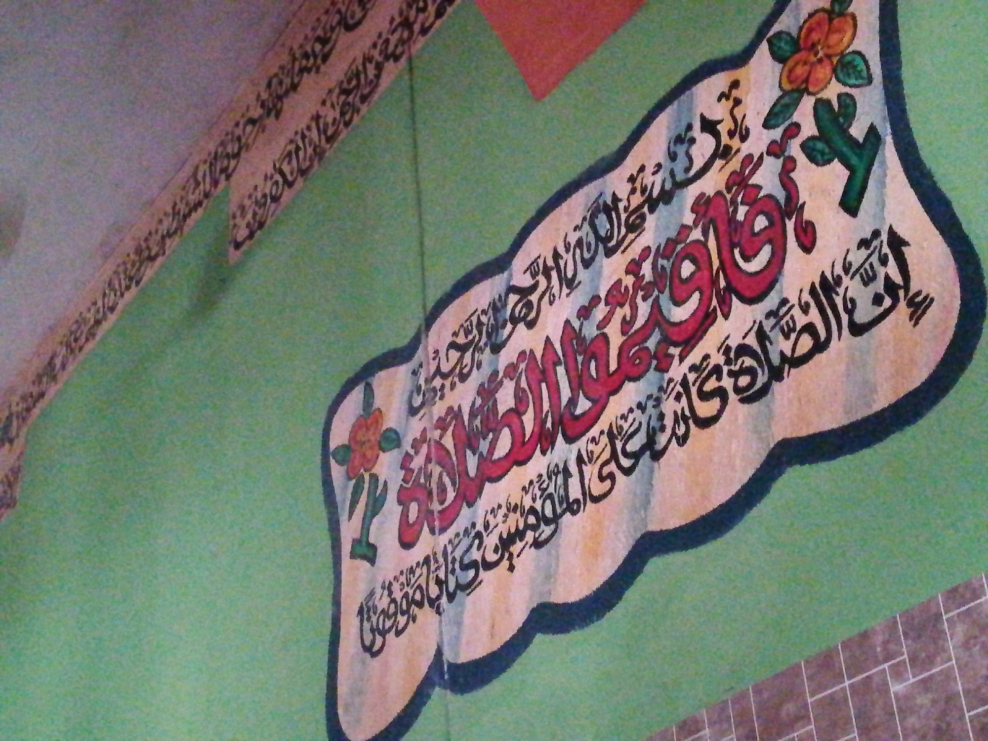The image shows a section of a wall in a building, possibly a mosque or religious institution. The wall is predominantly green and features decorative Arabic calligraphy. The calligraphy is presented in two locations: a horizontal strip near the ceiling and a large, ornate plaque-like design. The plaque has an irregular shape, possibly resembling a leaf or stylized ornament, and is decorated with floral motifs. The calligraphy is primarily in black and red inks. The bottom right corner of the image shows part of a tiled floor.
The image is likely from Lagos, Nigeria.