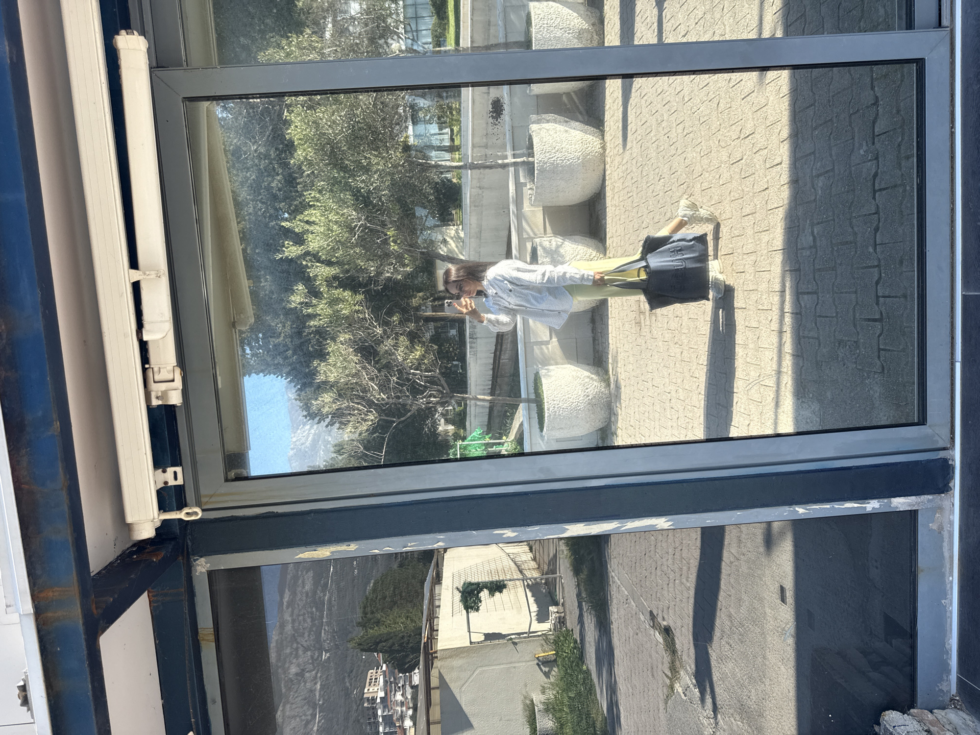 The image shows a reflection in a window, with a woman taking a photo of herself with her phone. She is wearing a white patterned shirt, green pants, and white sneakers, and has a black tote bag beside her. The reflection also captures a paved area with large, textured planters and trees. Outside the window, there is a view of buildings and greenery, possibly a cityscape nestled against a hill or mountain. The window frame is visible, with a light-colored blind or shade mechanism on the left side and some peeling paint around the frame.