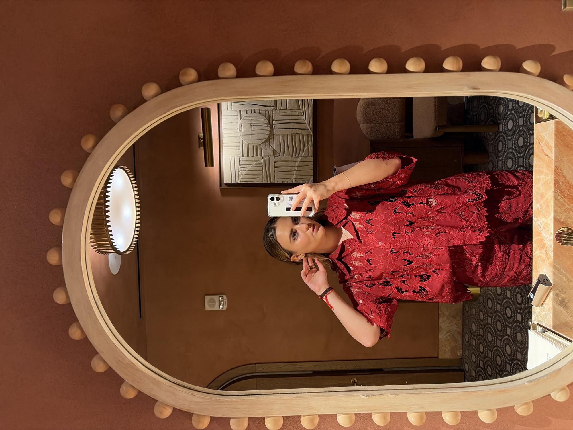 The image shows a woman in a red outfit taking a selfie in a mirror. The mirror has an oval shape and a decorative border of light wooden balls. The background reflected in the mirror reveals an interior space with artwork on the wall, a light fixture, a chair, and patterned tiles. The wall outside the mirror is a solid reddish-brown color. There is no information provided in the image to confirm the location. Therefore, the city or country in which the image was taken cannot be confirmed.