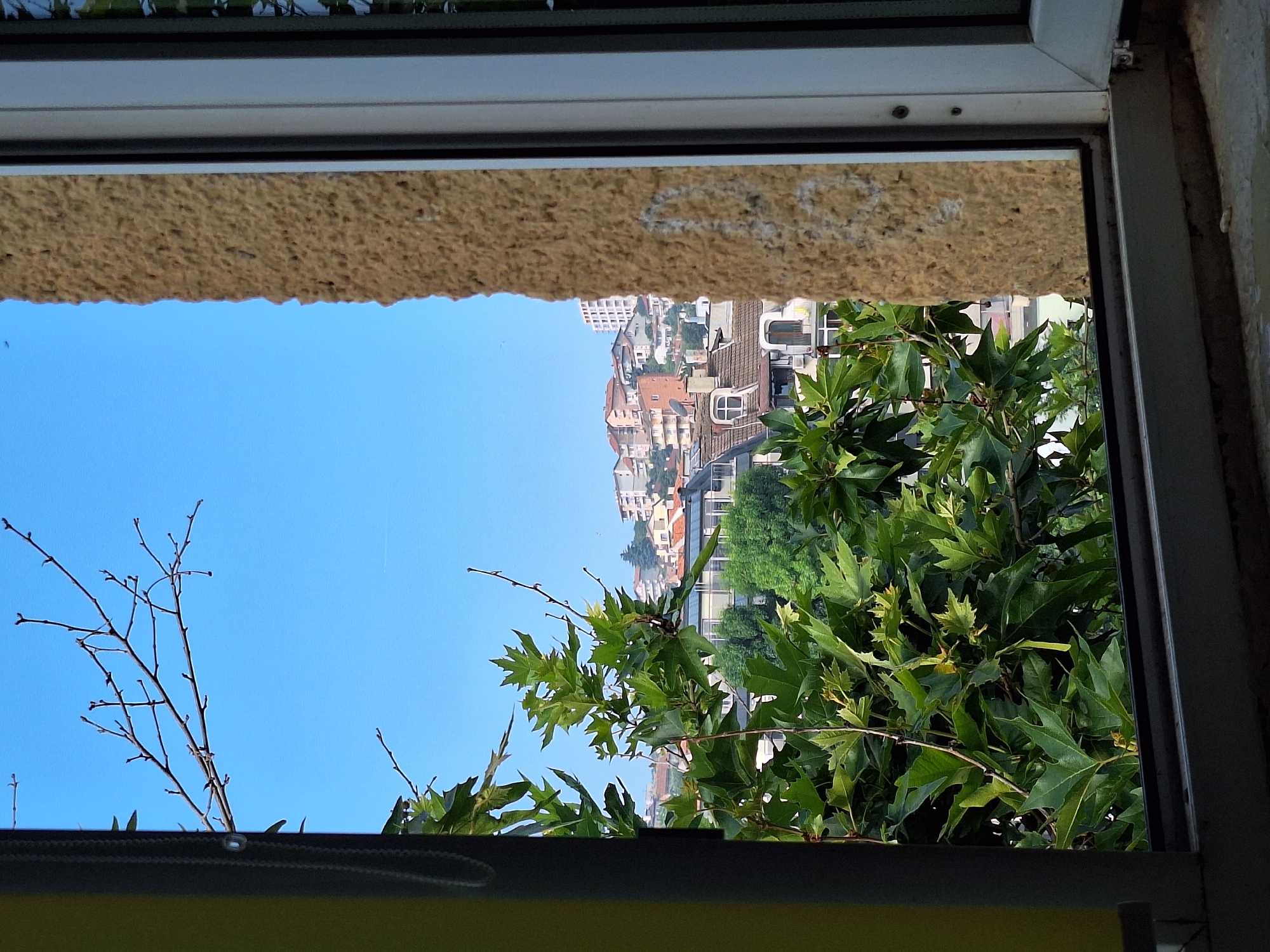 The image shows a view through a window frame in Varna, Bulgaria. In the foreground, to the left, is a portion of a yellow curtain and a dark grey window frame. A tree with large green leaves and bare branches partially obscures the view. To the right of the window frame is a rough, textured, light brown wall. Beyond the immediate foreground, the scene opens up to a cityscape. Buildings with orange and white roofs, some with satellite dishes, are visible on a hill under a clear blue sky. A tall, white modern building is also present in the distance. The overall impression is a residential area on a sunny day.