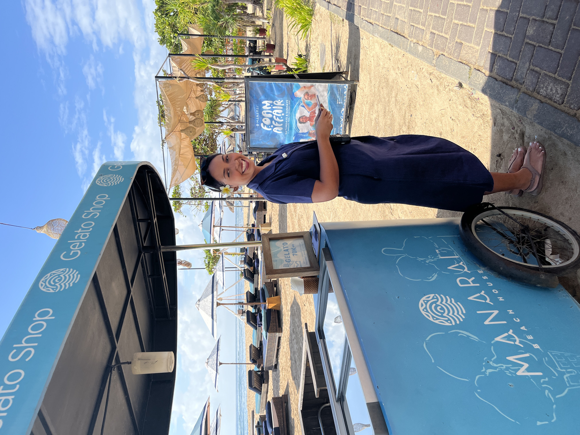 In Kuta Selatan, Indonesia, a woman stands on a sandy beach near a blue gelato cart. She is smiling at the camera and wearing a dark blue uniform dress and sandals. Behind her, beach chairs and umbrellas are arranged on the sand, and the ocean is visible in the distance. A sign advertising a "Foam Affair" event is propped up near the gelato cart. The scene is illuminated by daylight under a clear blue sky.