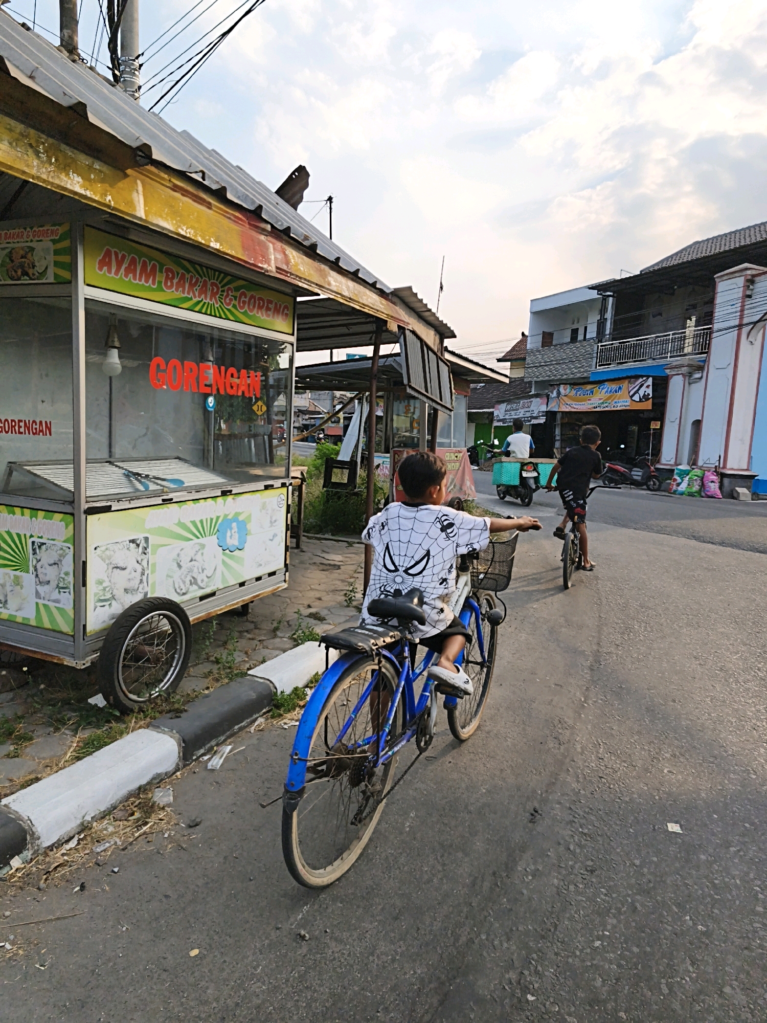 In Trayu, Indonesia, two young boys are riding bicycles on a paved street lined with small buildings. The boy in the foreground, wearing a white shirt with a Spider-Man design, is on a blue bicycle. His hand is extended forward, and he is looking ahead. Behind him, another boy on a black bicycle is pedaling away from the camera, heading towards the right side of the street. The street also has a food cart on the left side, displaying the words "AYAM BAKAR & GORENG" and "GORENGAN," indicating it sells grilled chicken and fried food. The sky is partly cloudy, and the scene appears to be during the late afternoon or early evening. The buildings on the right have multiple stories and balconies. Several motorbikes and bags of what appear to be fertilizer or grain are visible on the side of the road near the buildings.