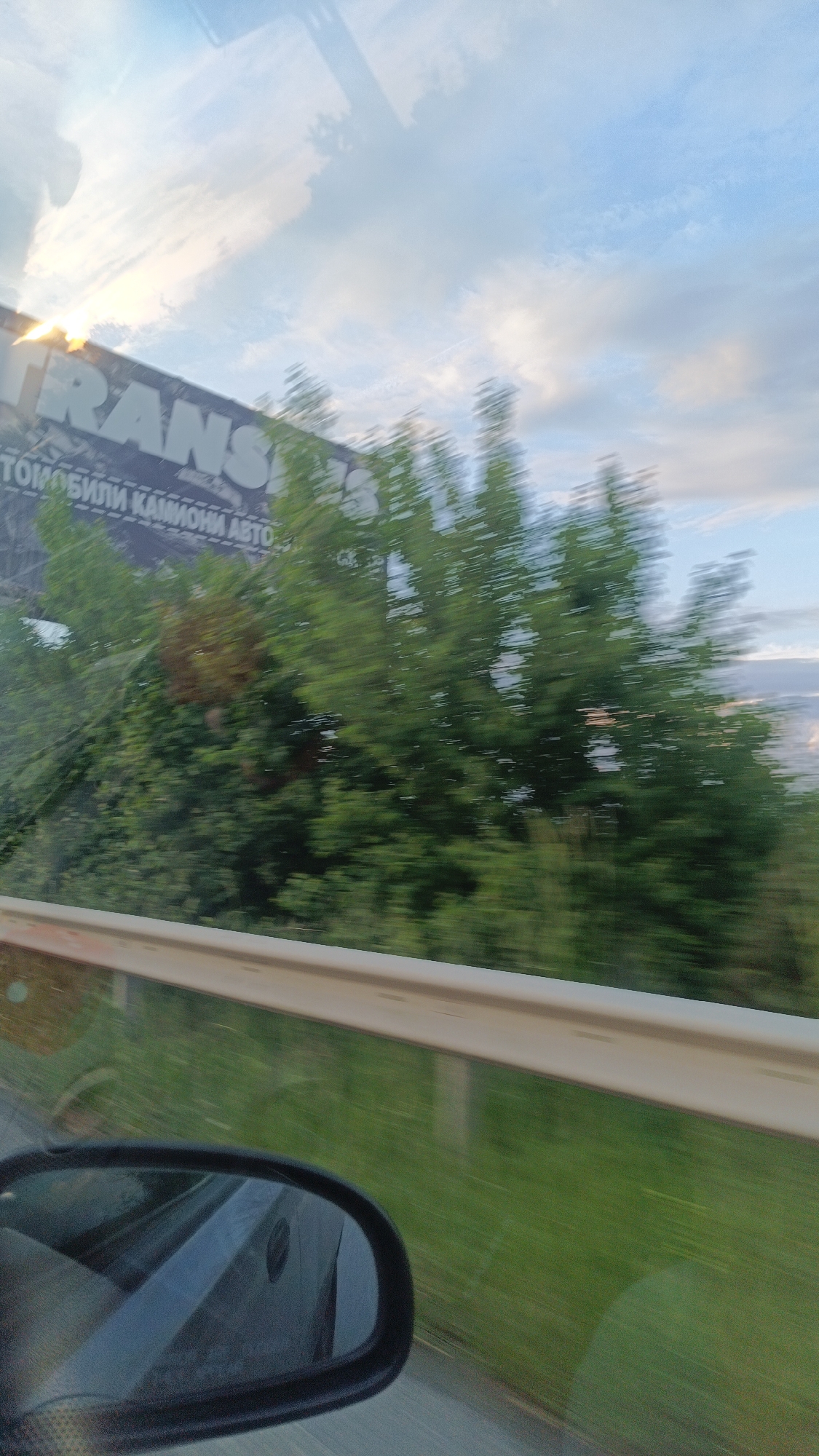 The photo, taken from inside a moving vehicle, shows a blurred roadside scene. A metal guardrail is visible along the right edge. Beyond it, grass blurs by. The car's side mirror is visible in the lower left. Through the windshield, trees partially obscure a sign with the word "TRANS" visible and text in Cyrillic, "TОМОБИЛИ КАМИОНИ АВТО". The sky is visible in the background with scattered clouds. Given the Cyrillic text, the location is likely in a Slavic-speaking country. The sign and guardrail indicate it is on a road outside Burgas, Bulgaria.
