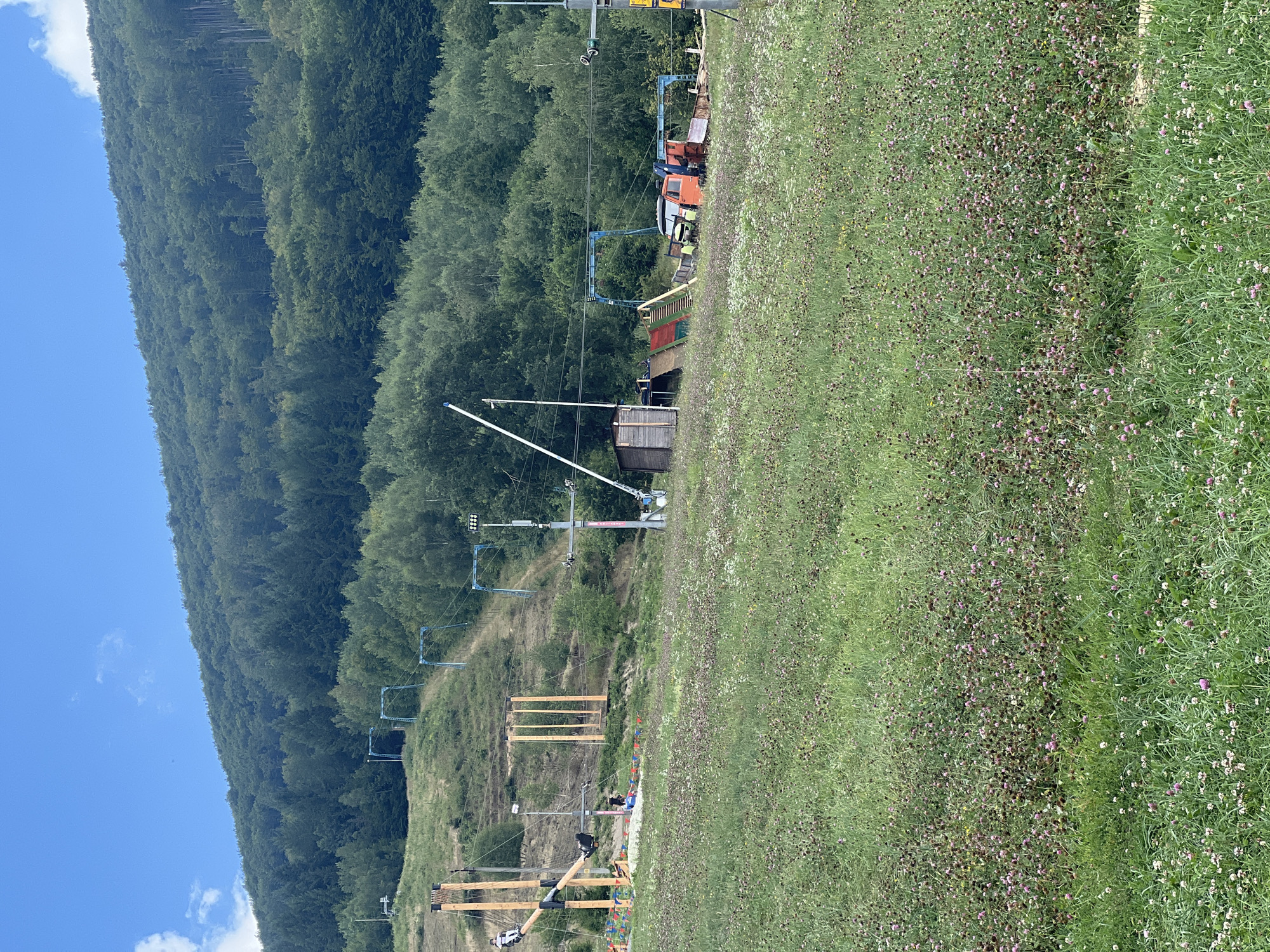 The image depicts an outdoor recreational area with a forest-covered hillside in the background. In the foreground, a field of green grass is dotted with small pink and white flowers. Further up the slope, there are various structures and activities associated with a summer sports or adventure park. This includes what appears to be a zipline or rope course with wooden platforms and cables, as well as ski lift equipment repurposed for summer use. Several people are visible, some engaged in activities on the structures, while others are walking around. Two colorful vehicles, possibly a small tractor and a larger orange truck, are parked near a small wooden building and a ramp. The sky is blue with some scattered white clouds. The overall scene suggests a sunny day at a resort or park offering a mix of nature and adventure activities. The location is identified as Сонячне, Ukraine.