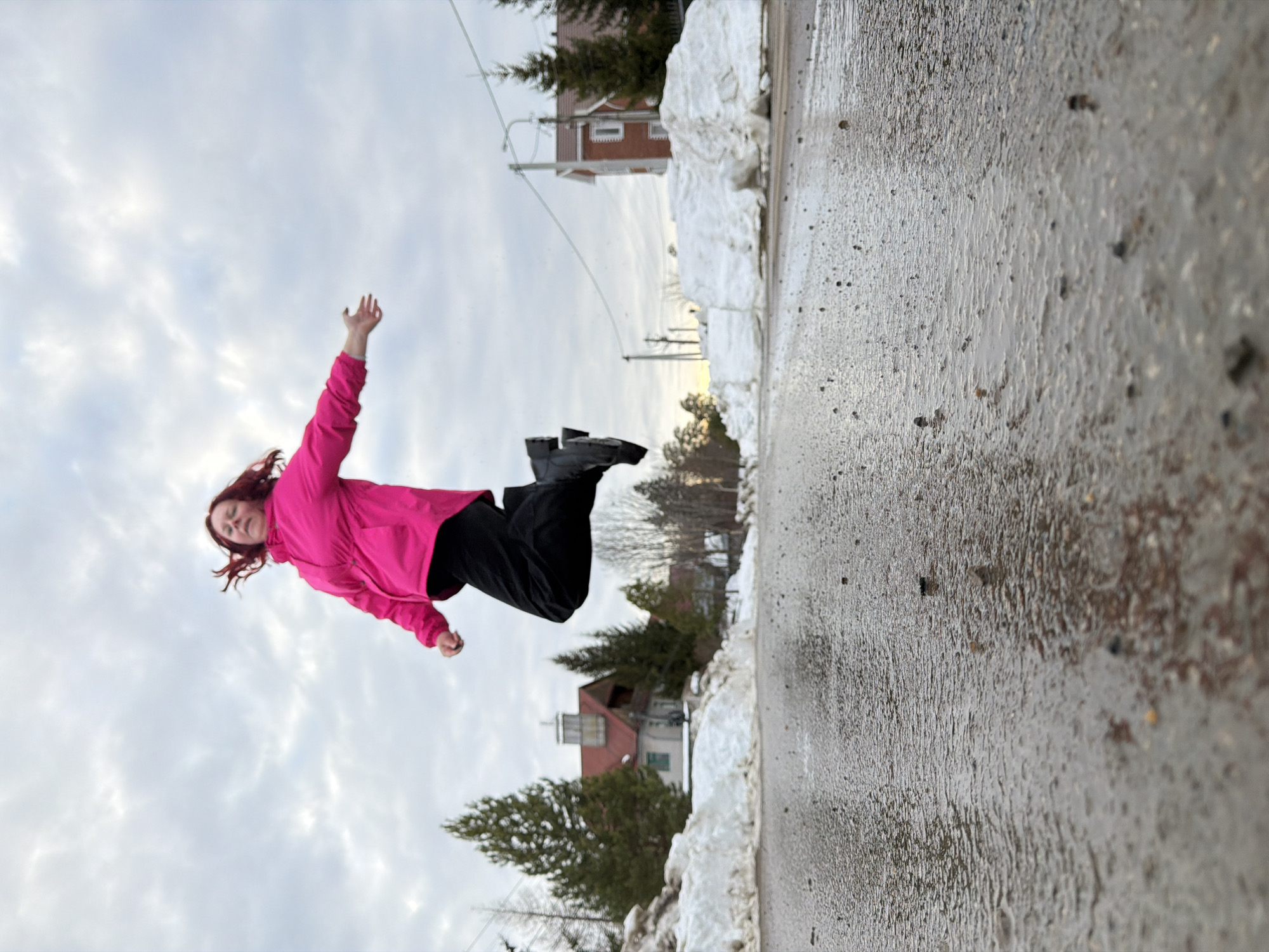 The image displays a single female subject in mid-air over a wet, paved road in a residential or semi-rural area of Chaikovsky, Russia. The individual, with reddish-brown hair, wears a bright pink jacket, black pants, and dark boots; her legs are bent, and her right arm is extended outwards. The foreground presents a low-angle view of a damp asphalt surface with small stones and visible puddles. Large, melting snow piles line both sides of the road in the midground. In the background, evergreen trees are interspersed with buildings. To the left, a multi-story house with a red-tiled roof and a cupola is partially visible. To the right, another red-roofed house with light-colored walls stands further back. The sky is overcast with a uniform layer of grey clouds. Utility poles and power lines extend across the mid-background.
