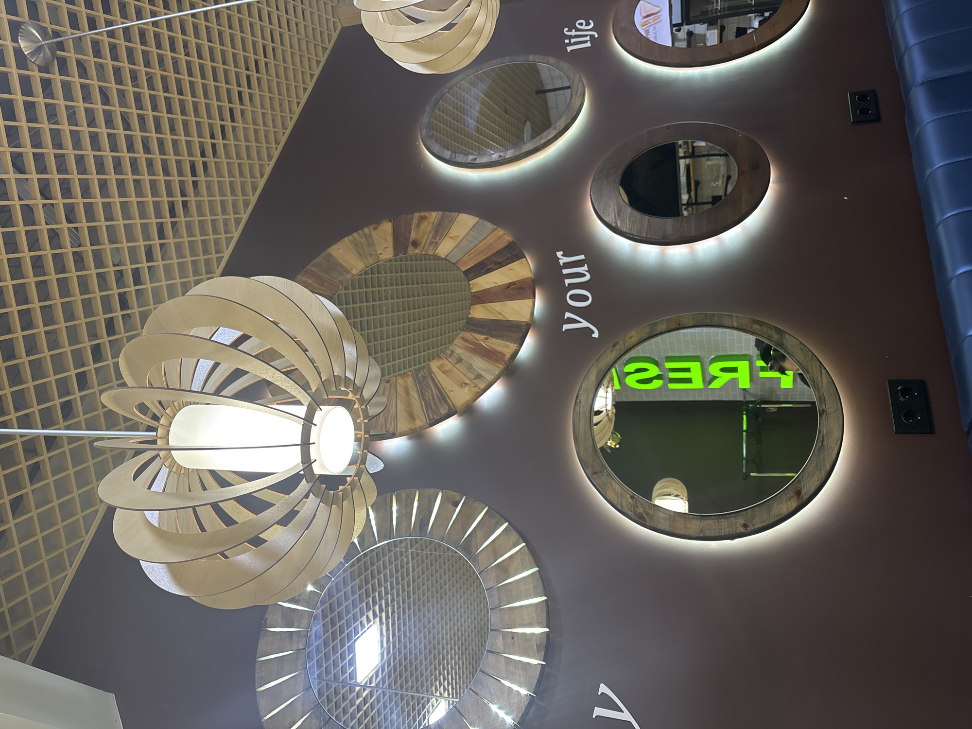 This is a photograph of a wall decorated with multiple round mirrors, wooden lamps, and text. The mirrors have rustic wooden frames and are illuminated from behind, creating a soft glow. Some mirrors reflect a ceiling with a grid pattern and lighting fixtures. One mirror reflects green neon lettering that spells out "FRESH" in reverse. Above the mirrors, a wooden-patterned ceiling with a grid design is visible. The wall itself is a dark brown color. The text "your" and "life" are mounted on the wall in white, stylized lettering, suggesting a phrase related to freshness or well-being. In the lower part of the image, a dark blue upholstered bench is visible, and a black electrical outlet is mounted on the wall. The scene appears to be an interior space, possibly a cafe or a retail establishment. The presence of the "FRESH" lettering and the overall aesthetic suggest a focus on health, food, or a welcoming atmosphere. Without further information, it is impossible to determine the specific location in Naberezhnye Chelny, Russia, or identify any individuals present.