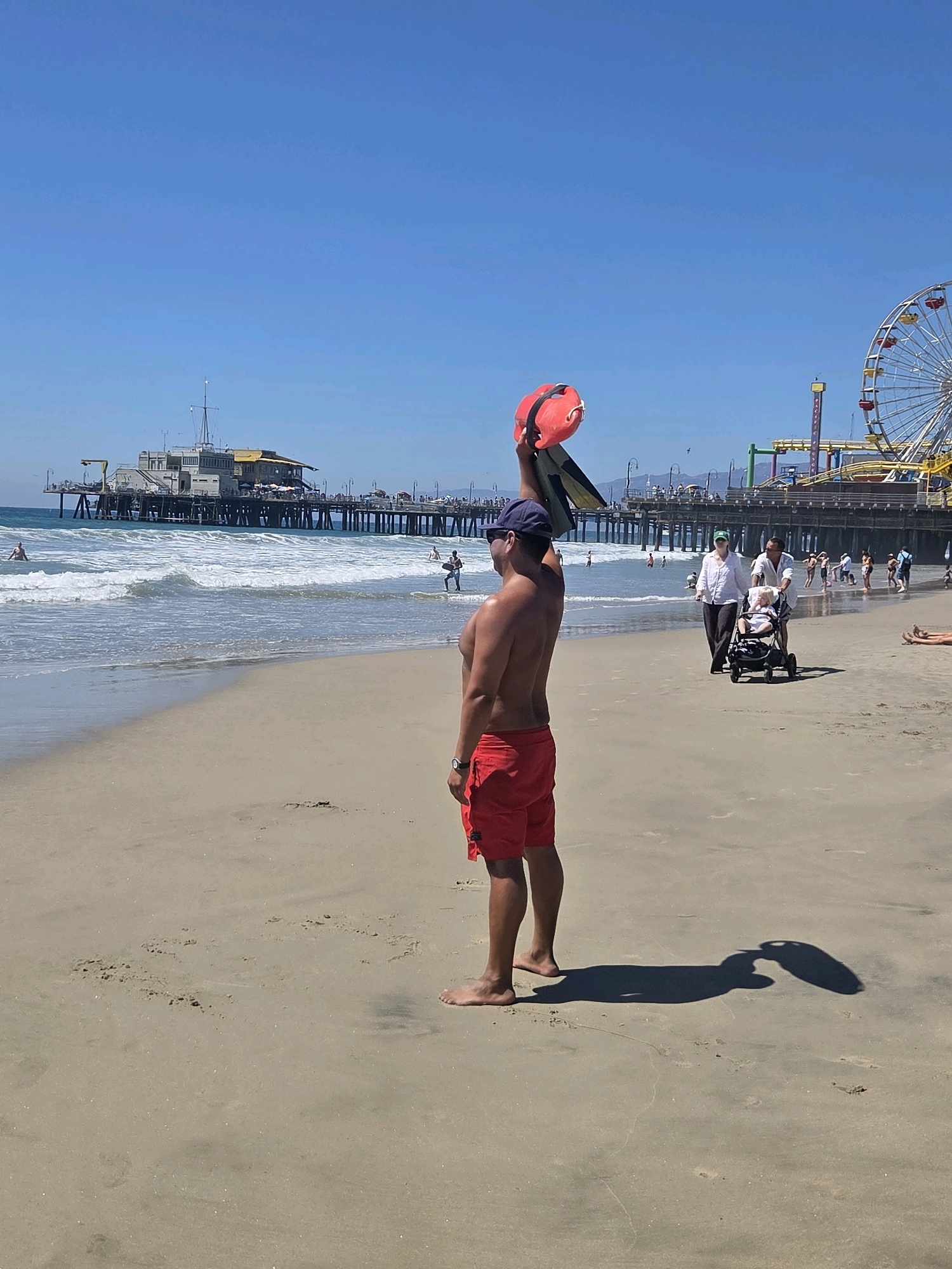 A man stands on the sand at Santa Monica Beach, facing away from the camera. He wears red swim trunks and a blue baseball cap. He holds a red and black flotation device above his head. His shadow stretches across the sand in front of him. In the background, waves break on the shore and people walk along the water's edge. A pier with buildings and a Ferris wheel is visible in the distance. The sky is clear and blue.