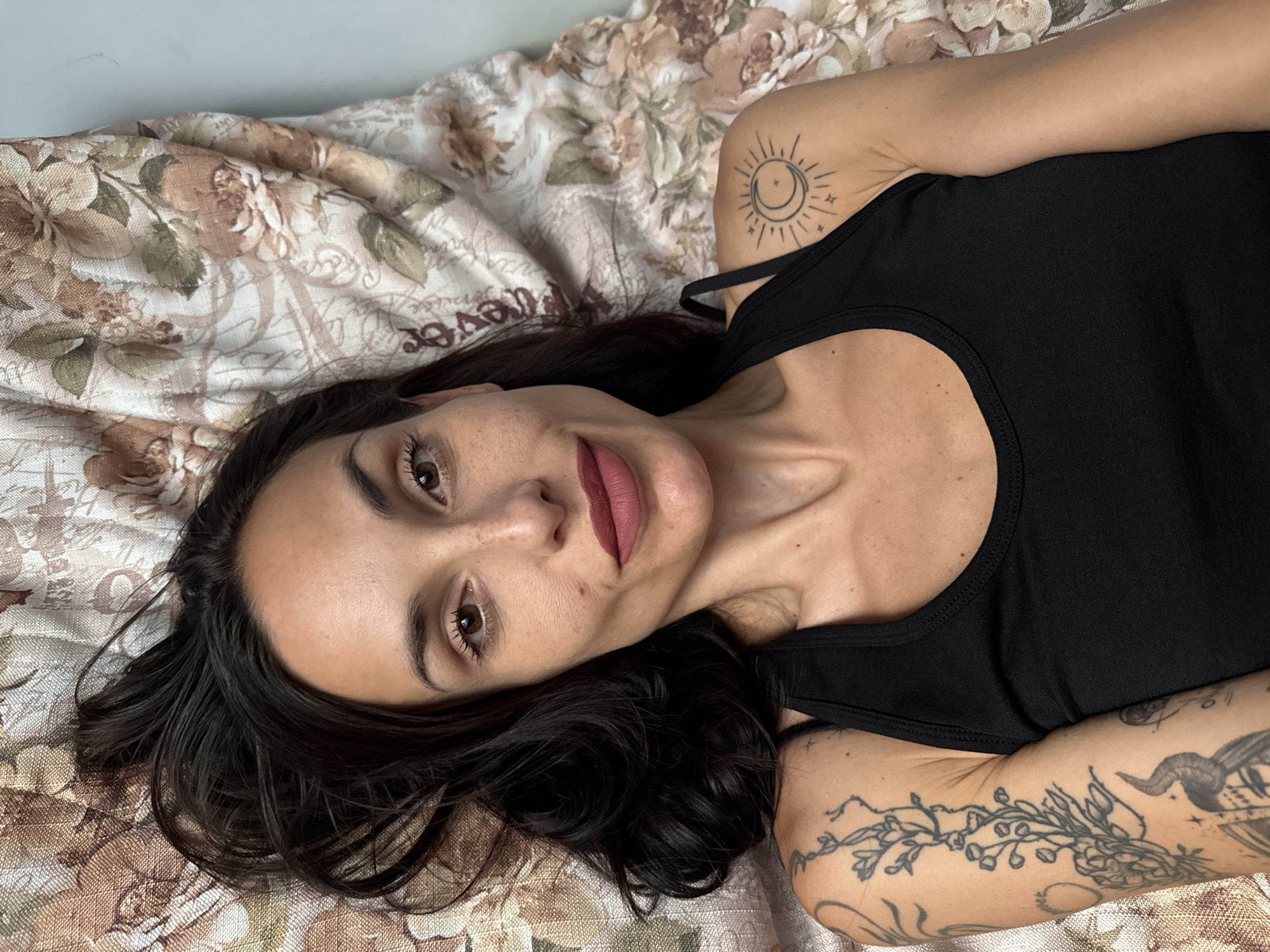 A woman with dark hair, wearing a black tank top, is lying on a bed with a floral-patterned bedspread. She is looking directly at the camera. She has a sun and moon tattoo on her left shoulder and floral and other tattoos on her right arm. The location is not discernible as being in Pleven, Bulgaria based on the available visual information.