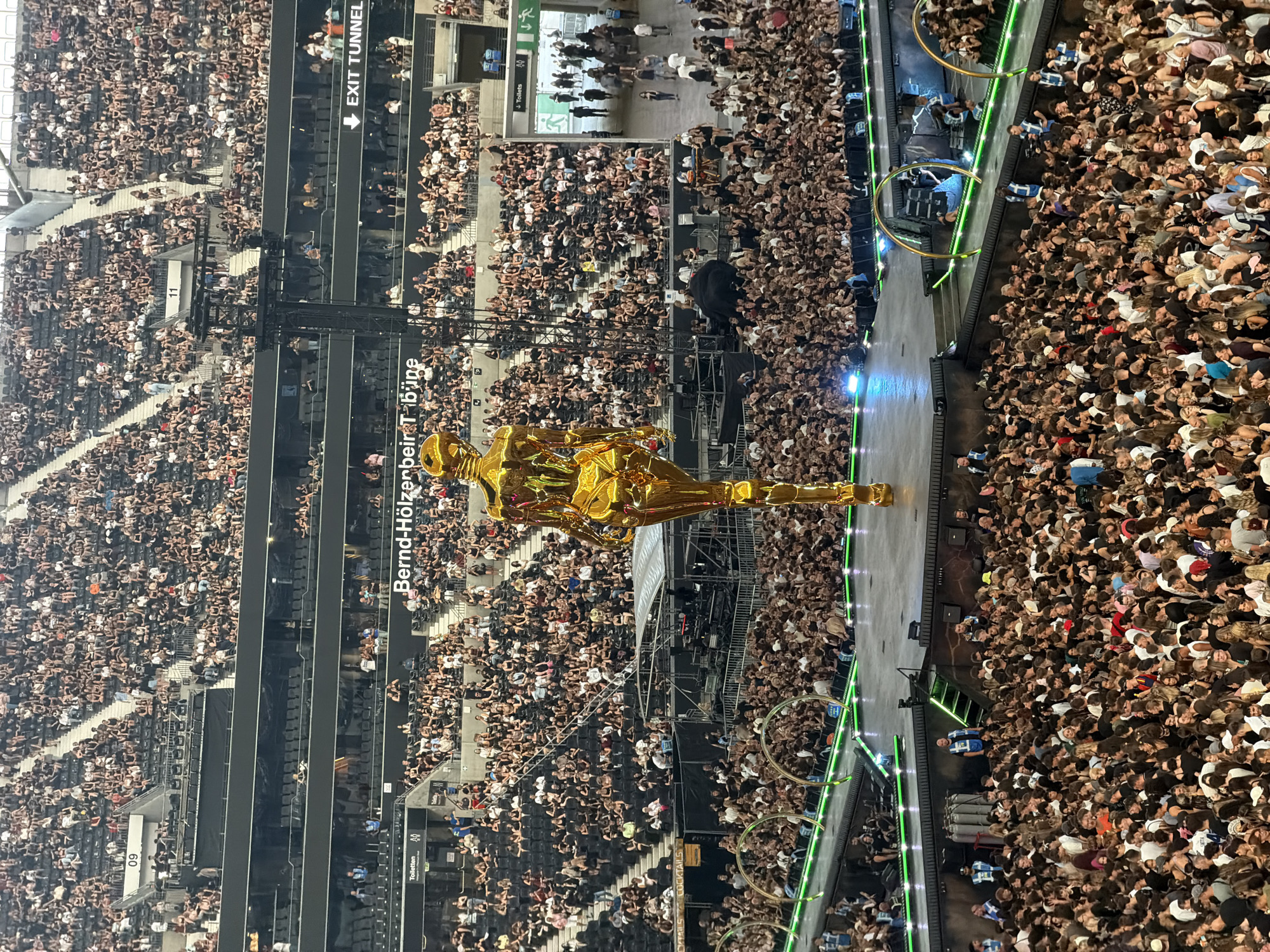 The image depicts a large indoor concert or event venue filled with a dense crowd of spectators. In the foreground, a towering, golden, humanoid sculpture is prominently displayed on a raised stage. The sculpture has a stylized, elongated form with smooth, reflective surfaces. The stadium seating, visible in the background and surrounding the audience, is largely empty in the upper sections, suggesting the event is concentrated in the lower bowl. The audience members are diverse in age and gender, predominantly dressed in casual attire. Many are looking towards the stage, with some holding up phones, indicating they are recording or photographing the event. The environment appears to be well-lit, with stage lighting evident in the lower portion of the image. Signs are visible on the stadium structure, including "09" and "11" on numbered sections, and an "EXIT TUNNEL" sign. Another sign reads "Bernd-Holzenbein-Tribune," which is a section of the Deutsche Bank Park stadium in Frankfurt, Germany, known for hosting major sporting events and concerts. The presence of this sign confirms the location. The overall scene conveys a sense of a large-scale public gathering, likely a concert or a similar entertainment event, with the golden sculpture serving as a central visual element.