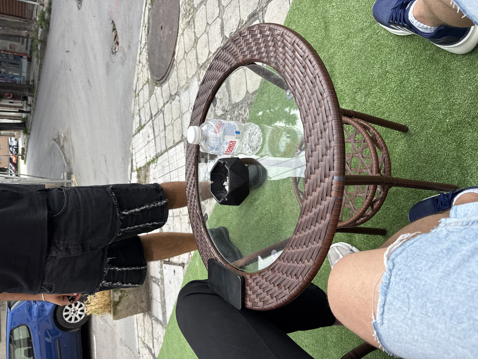 The image captures an overhead view of two people seated at an outdoor cafe in what appears to be Varna, Bulgaria, during the daytime. The perspective looks down onto a round table with a glass top and a brown, woven wicker-style frame.
On the table, a plastic bottle of water is prominently displayed, bearing the visible text "RIBANIA" and "ACQUA MINERALE NATURALE". Beside it rests a black Moka pot or similar coffee maker. A black smartphone is also placed on the table, near the edge.
The ground around the table and chairs is covered with artificial green grass, contrasting with the light-colored, patterned cobblestone pavement that extends into a concrete street.
Two individuals are partially visible:
- To the left, one person wears black distressed shorts and black sneakers, with bare legs. A blue car is parked on the street in the background.
- To the right, another person wears light blue denim shorts and navy blue sneakers with white soles and laces. White socks are also visible.
The setting suggests a casual outdoor seating area next to a public street, under bright, clear daylight. The activity appears to be simply sitting and enjoying the outdoor environment, perhaps having a drink. The overall scene is bright and conveys a relaxed, urban atmosphere.