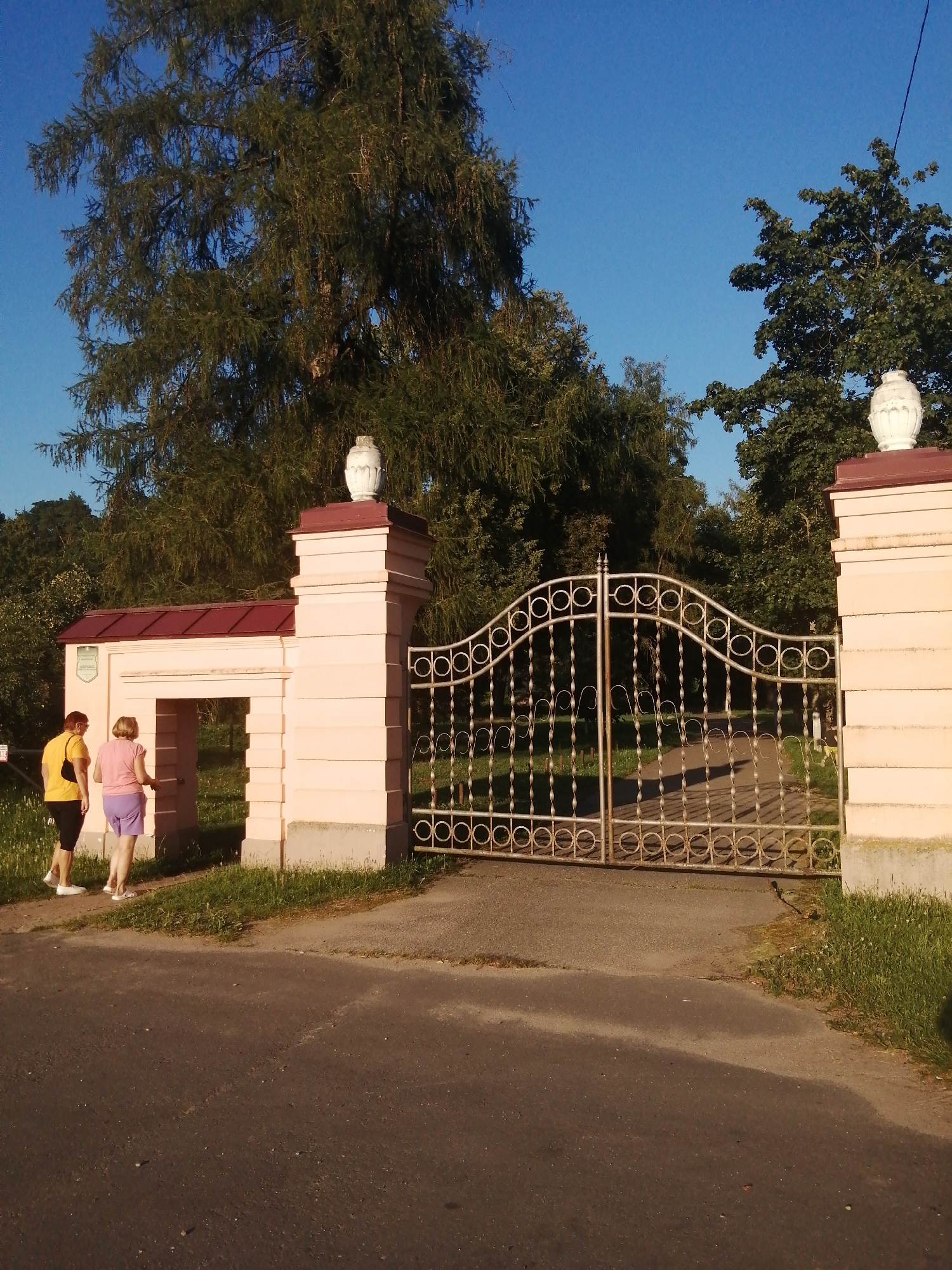 In Slonim, Belarus, two women stand near an ornate gate at a park entrance. The woman on the left wears a yellow t-shirt and black shorts, while the woman on the right is in a pink t-shirt and purple shorts. They are positioned at the archway of a pink stucco structure with a red tiled roof. The gate itself is made of decorative metalwork with circular and spiral patterns. Beyond the gate, a paved path leads into a wooded area. The scene is illuminated by clear blue sky, suggesting a sunny day.