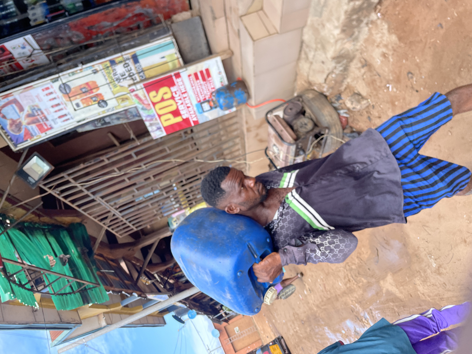 A man in blue striped shorts and a patterned shirt carries a large blue plastic container on his shoulder in Onitsha, Nigeria. He is standing on a dirt road. Behind him, there is a shop with various signs, including one that says "POS Service Available Here." There is a generator next to the shop.