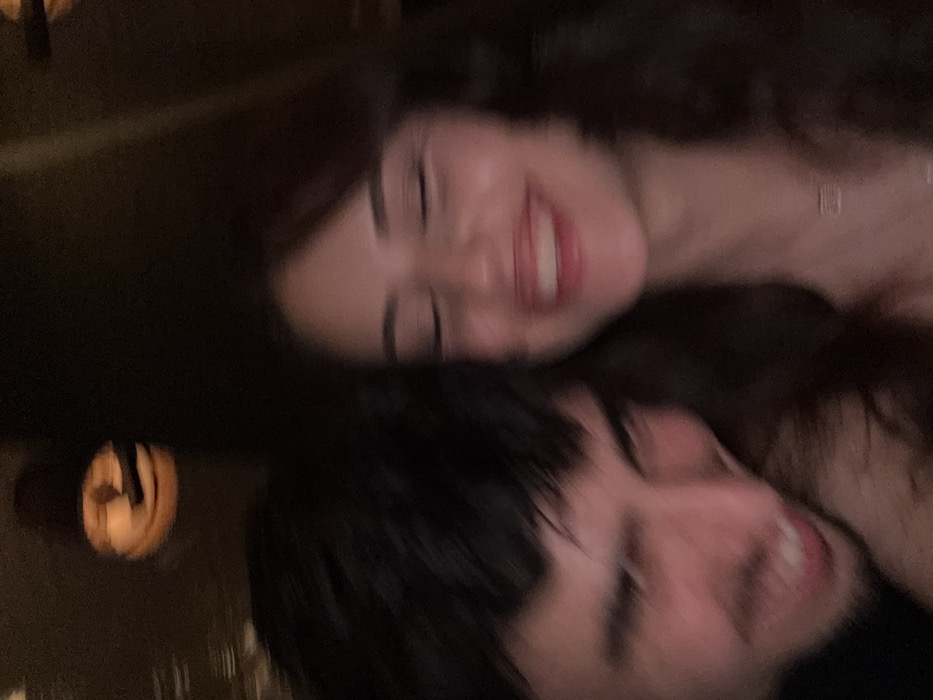 The image captures a blurry, close-up shot of two individuals, likely a couple or close friends, sharing a joyful moment. The photo is rotated, making the subjects appear to be lying on their sides.
The person at the top has long, dark, wavy hair and a fair complexion. They are smiling broadly with their eyes squinted or closed in an expression of happiness or laughter, revealing their teeth. A faint light-colored necklace or top strap is visible around their neck.
Beneath and slightly behind them is the second person, whose face is partially obscured. This individual has short, dark hair and also appears to be smiling widely, showing teeth, with their eyes similarly squinted or closed.
The overall setting is dark and dimly lit, suggesting an indoor environment, possibly at night. The background is out of focus, enhancing the intimacy of the moment. There are blurry, warm-toned light sources visible in the dark background, appearing as soft orange or yellow glows, possibly from lamps or decorative lighting. The blurriness of the image, potentially due to motion or low light conditions, adds to a candid and dreamlike quality, emphasizing the shared emotion of happiness and affection between the two individuals. No text is visible in the image.