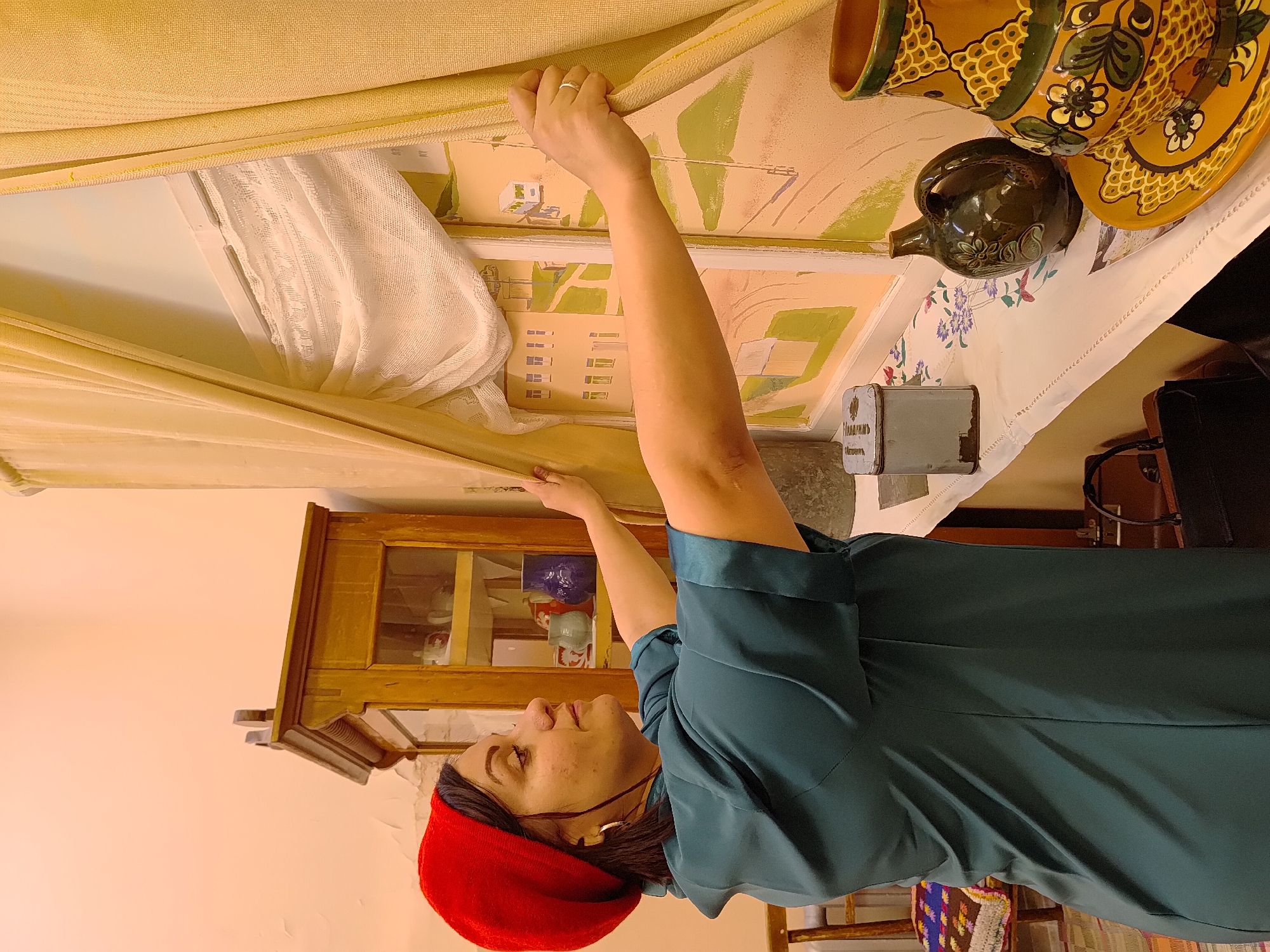 An indoor scene, likely in Markovsky, Russia, shows a woman in profile engaged in an everyday activity. She wears a red beret and a dark teal-colored top or dress, with her right arm raised, adjusting or holding back a light-yellow curtain. Behind this main curtain, a sheer white lace curtain is visible, and further back, a wall features painted murals depicting stylized green landscapes or architectural elements.
On a table covered with a white tablecloth with embroidered edges, a collection of ceramic ware is displayed. This includes a large yellow and green ornate jug and a matching plate or bowl, alongside a smaller dark green pitcher with similar floral decorations. A grey metal box with the Cyrillillic inscription "Марковский Почтамтъ" (Markovsky Post Office) rests on the tablecloth, providing a direct location cue. In the background, a wooden display cabinet with glass doors can be partially seen. The woman's gaze is directed upwards, suggesting focus on the curtain she is handling. The overall setting evokes a sense of traditional or cultural ambiance. The lighting is indicative of an indoor daytime scene.