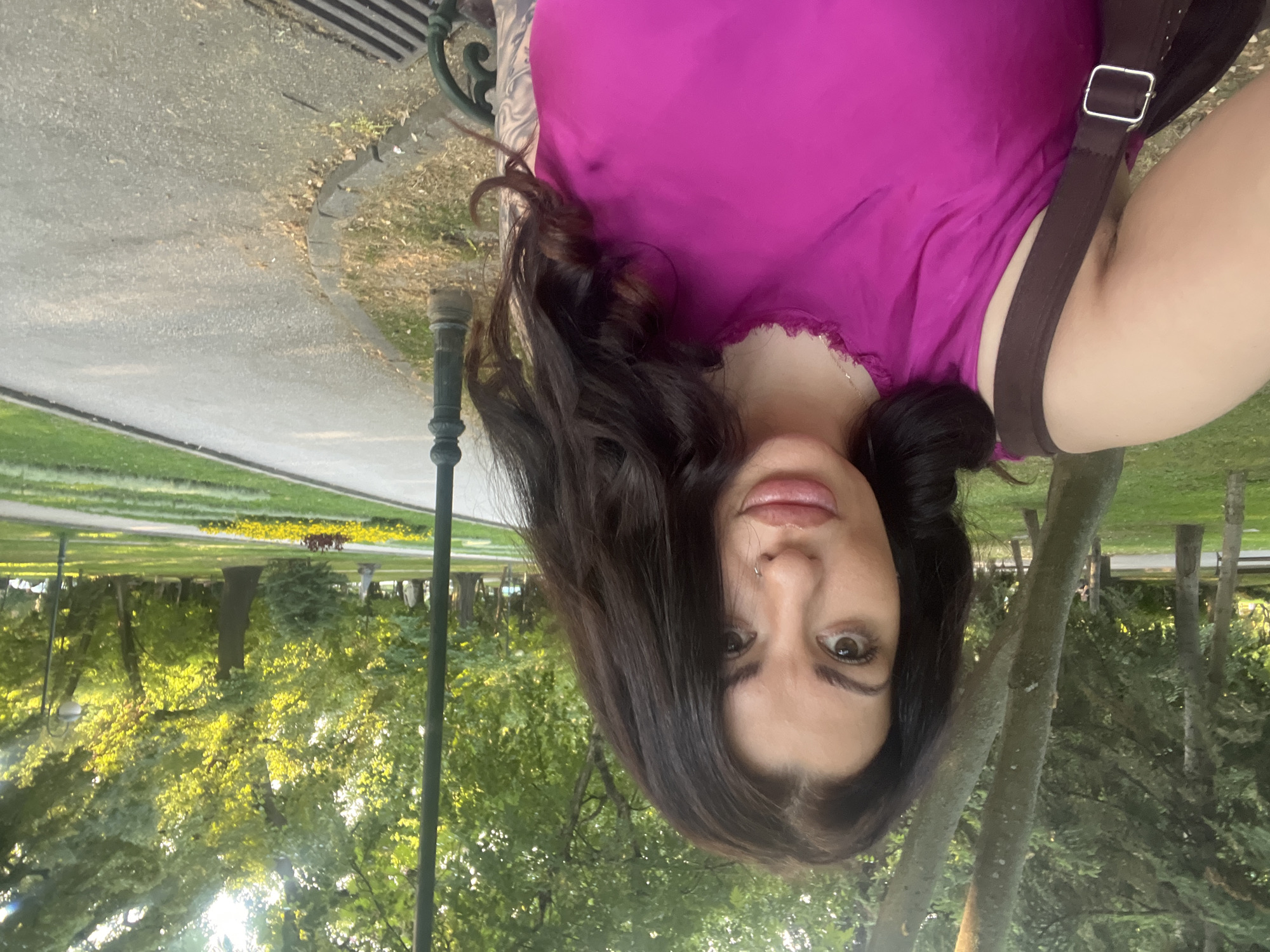 A woman with dark, curly hair and a nose ring takes a selfie in a park. She is wearing a bright pink, sleeveless top and a brown strap from a bag is visible over her shoulder. The background shows a path winding through a lush green park with trees and flowerbeds. The setting appears to be Borisova Gradina in Plovdiv, Bulgaria, characterized by its mature trees, manicured lawns, and paved walkways, suggesting a public recreational space. The lighting indicates it's late afternoon or early evening.