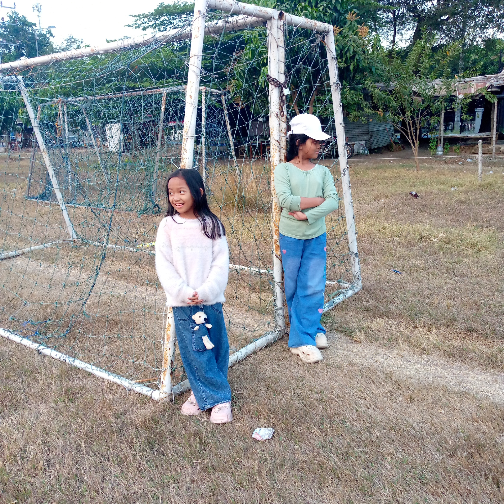 In Pacing, Indonesia, two young girls stand near a weathered soccer goal. The girl on the left, wearing a fluffy white sweater and blue jeans adorned with a teddy bear keychain, looks to her left with a smile. The girl on the right, sporting a light green long-sleeved shirt and blue jeans with pink heart accents, wears a white baseball cap and has her arms crossed. The soccer goal, made of rusty white metal and draped with a blue net, shows signs of age and wear. The ground around them is covered in dry, brown grass. In the background, trees and a rustic structure with a corrugated metal roof are visible, suggesting a rural or semi-rural setting. The overall scene captures a moment of childhood by a sports field.