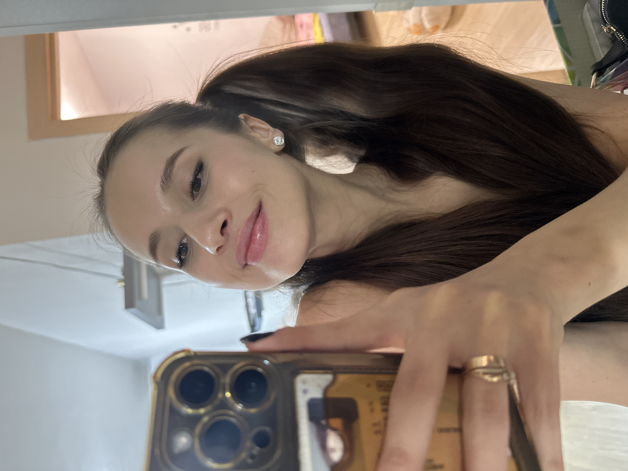 A woman with long, dark hair in a high ponytail is taking a selfie with a smartphone. She has fair skin, dark eyes, and full lips with a glossy sheen. She is wearing stud earrings. Her left hand is holding the phone, and a gold ring is visible on her ring finger. The phone has a gold-colored case and multiple camera lenses. The background is a brightly lit interior space with white walls and a framed mirror or picture. No specific details indicate the location in Yekaterinburg, Russia.