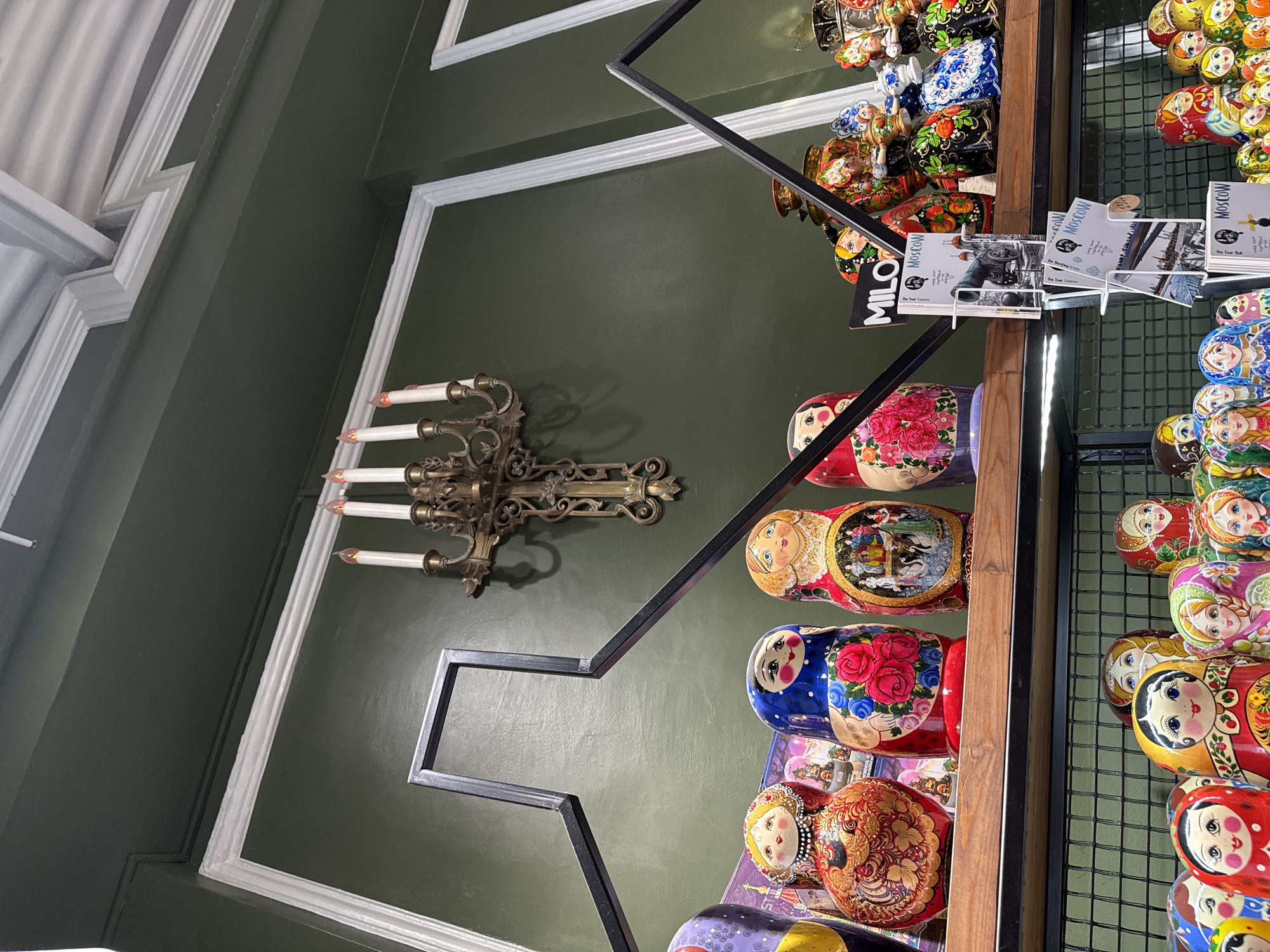 An indoor, eye-level shot depicts a collection of Russian nesting dolls displayed on shelves against a dark green wall. A vintage-style candelabra with five electric candles is mounted on the wall above the dolls. The dolls are varied in size and design, featuring colorful hand-painted patterns and faces. Some dolls are partially obscured by a black metal frame that appears to be part of the shelving unit. The shelves themselves are wooden, with a black metal grid below. A few postcards featuring images of Moscow are displayed in a white holder on the shelf. The lighting is somewhat dim, highlighting the details of the dolls and the candelabra.