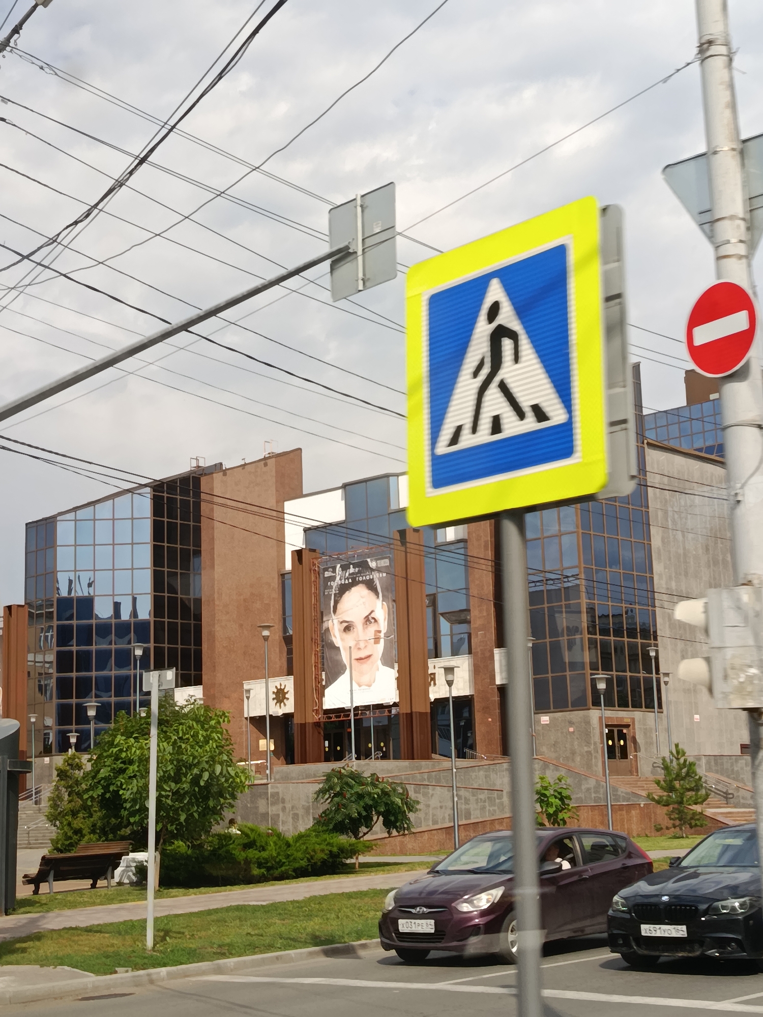The image shows a street scene in Saratov, Russia, on a cloudy day. In the foreground, a pedestrian crossing sign is prominently displayed, with a red circular "no entry" sign also visible on a pole to its right. Two cars, a dark purple hatchback and a black sedan, are parked or stopped at the intersection.
In the background, a modern building with a glass facade and brown accents stands. A large billboard featuring a woman's face is attached to the building's exterior. The building appears to be a commercial or public space, with multiple entrances and lampposts visible. A park bench and lush greenery are situated in front of the building, suggesting a public area. The overall impression is a typical urban environment with traffic and infrastructure elements. The sky is overcast, indicating a cloudy or possibly impending rainy day. No specific activity or event is clearly depicted beyond the presence of parked cars and pedestrian signage, implying normal street activity. Visible text includes license plates on the cars: "X031PE 64" and "X691YO 164". There is also some Cyrillic text on the billboard, but it is not clearly legible.