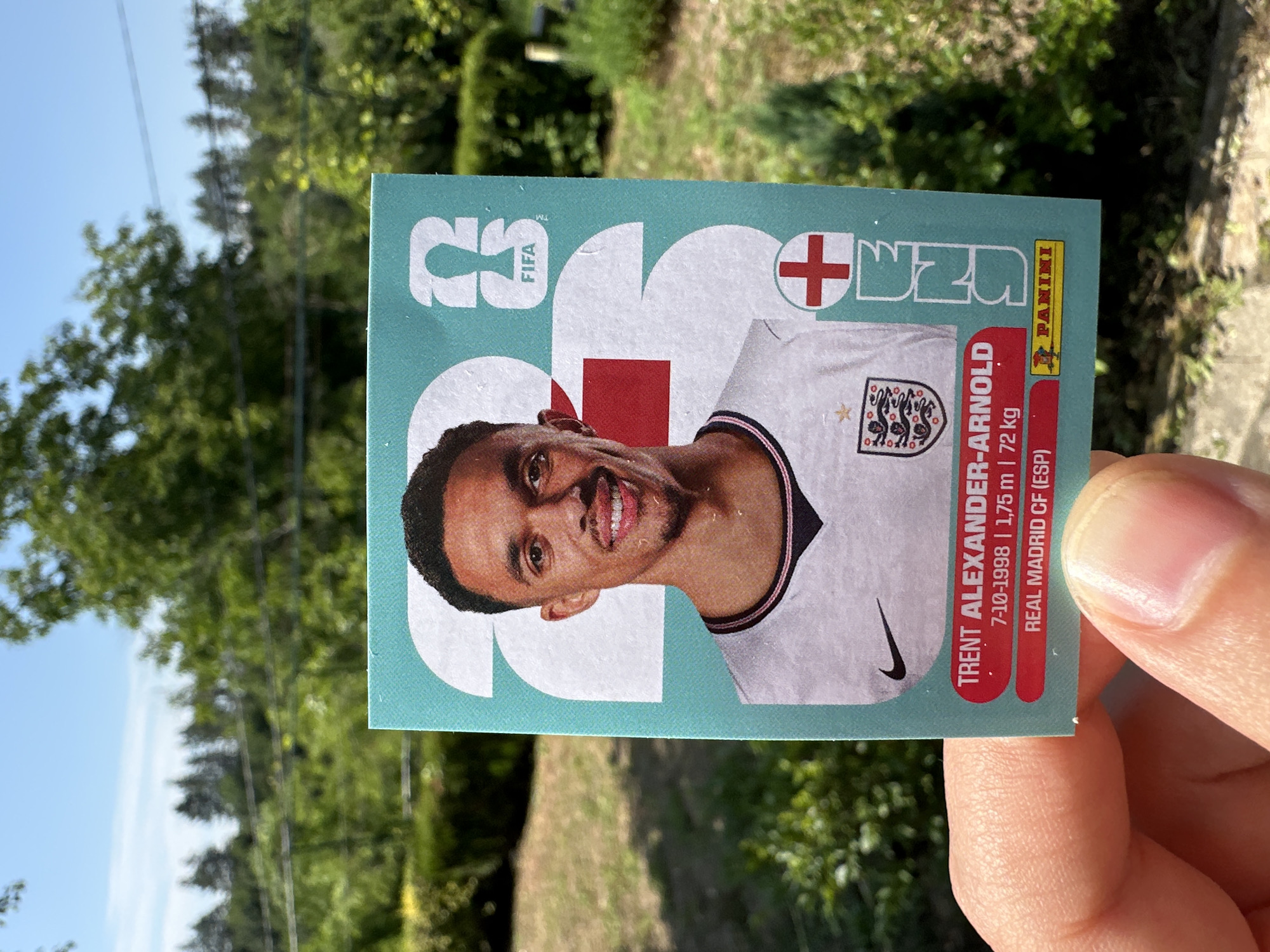 A person's hand holds up a Panini soccer sticker of Trent Alexander-Arnold in front of a blurred outdoor background. The sticker features a portrait of the player wearing an England jersey. Text on the sticker includes his name, birthdate, height, weight, and club affiliation (Real Madrid CF). The background consists of green trees and a clear blue sky, suggesting a sunny day. The location is identified as Prisovo, Bulgaria, based on the provided context.