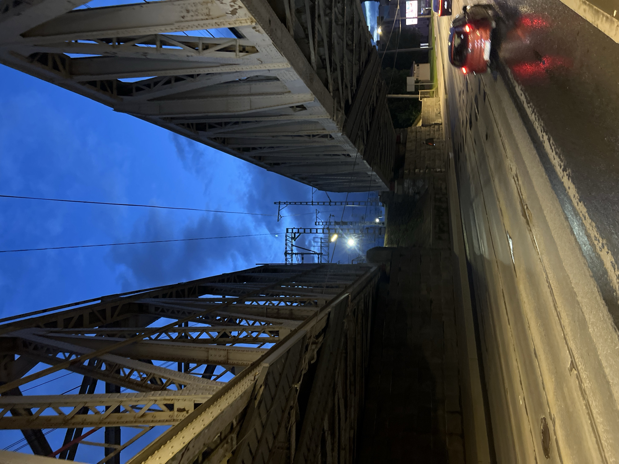 An outdoor, low-angle, eye-level shot captures the underbelly of two large, steel truss railway bridges meeting in the distance. The sky above is a deep twilight blue, with scattered clouds, and overhead railway lines and poles are visible. The bridge structures are predominantly grey, with visible rivets and a somewhat weathered appearance. The ground below is a wet, asphalt road with tram tracks, reflecting the lights from streetlamps and passing cars. The road surface is uneven, with some patches of standing water. A red car with its brake lights illuminated is visible in the right lane, moving away from the viewer. The scene appears to be an urban environment, possibly in Saint Petersburg, Russia, given the distinctive architecture of the bridges and the presence of tram tracks. The lighting suggests dusk or early evening.